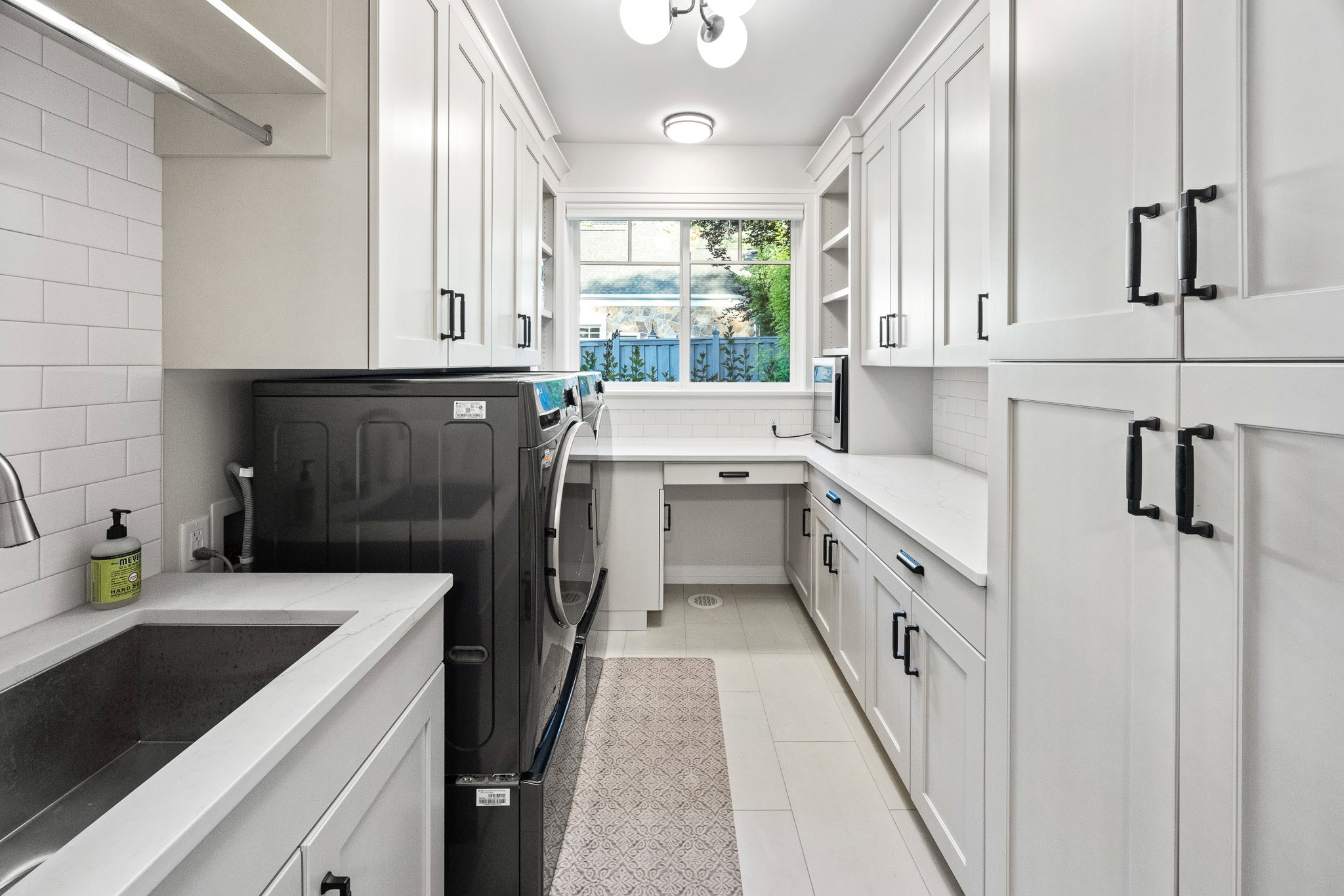 Bright utility room featuring white shaker-style cabinetry, a built-in workstation, undermount sink, and a window providing natural light