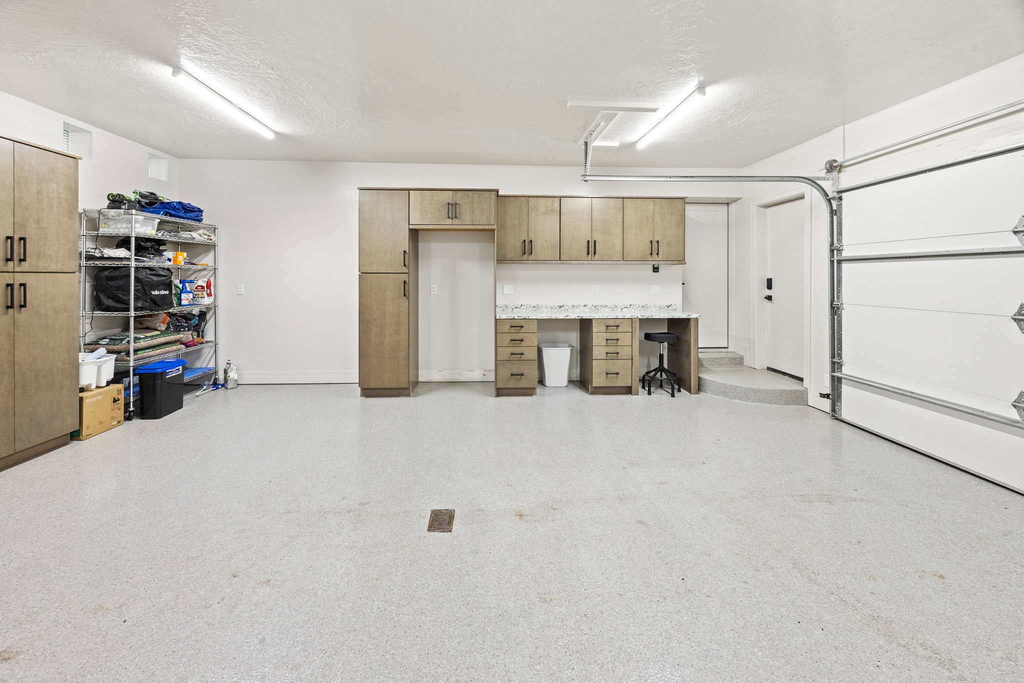 Spacious garage featuring a light-toned epoxy floor, extensive built-in cabinetry, an integrated workbench with a stone-finish countertop, and bright overhead strip lighting