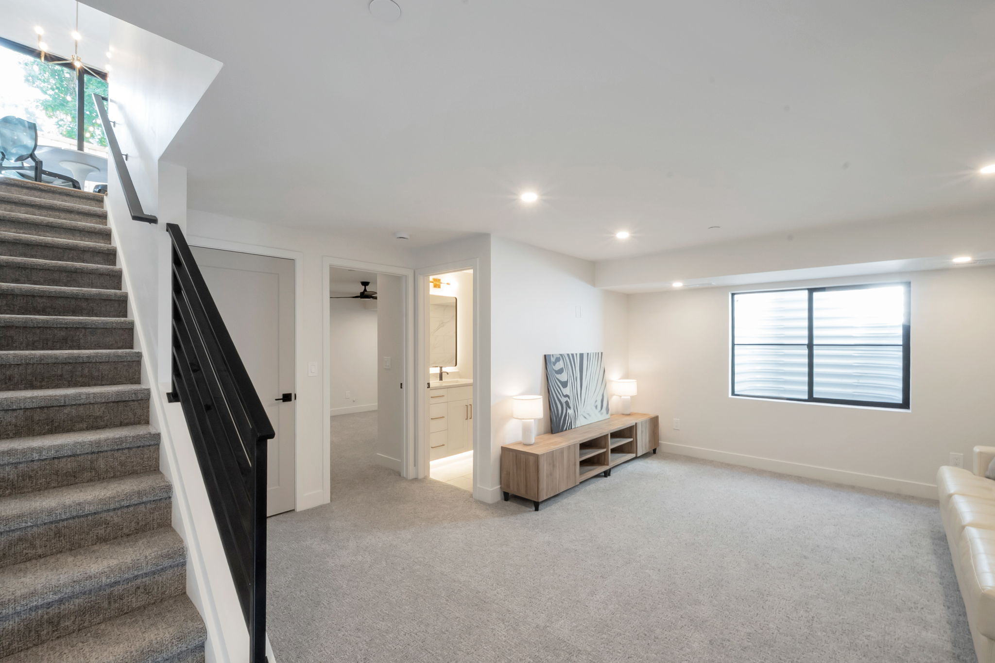 Carpeted staircase with black metal railing, leading to a spacious room with recessed lighting and a large egress window