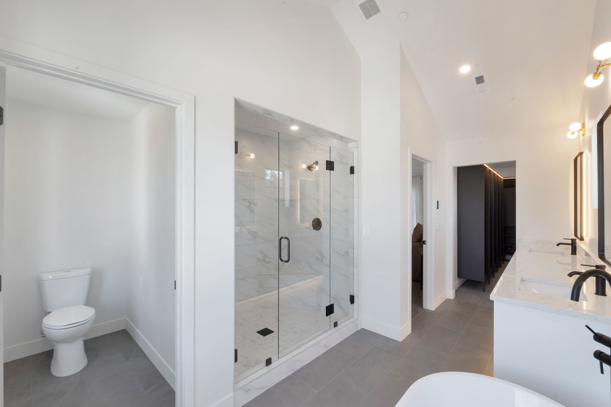 Spacious bathroom featuring a glass-enclosed shower with tile surround, dual vanity with undermount sinks and dark fixtures, and light gray floor tiles