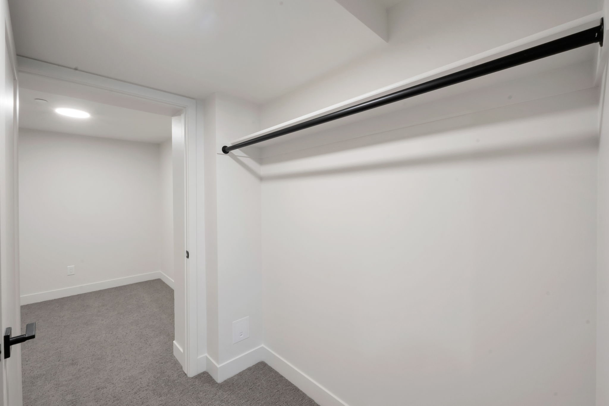 Interior closet space featuring a black hanging rod, white walls, and a light gray carpeted floor extending into an adjacent room with a recessed ceiling light