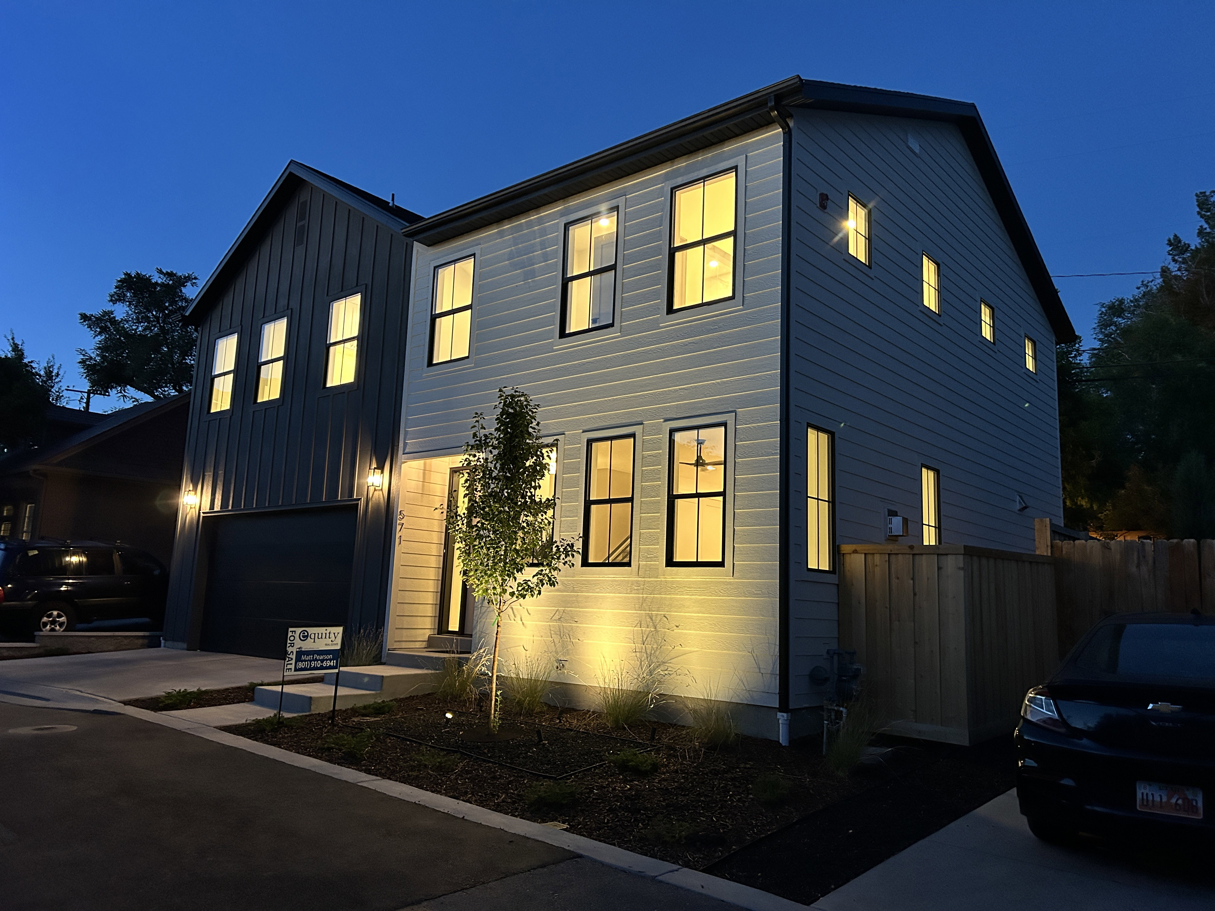 Modern architecture featuring vertical and horizontal siding, multiple windows with dark frames, a dark garage door, and exterior lighting