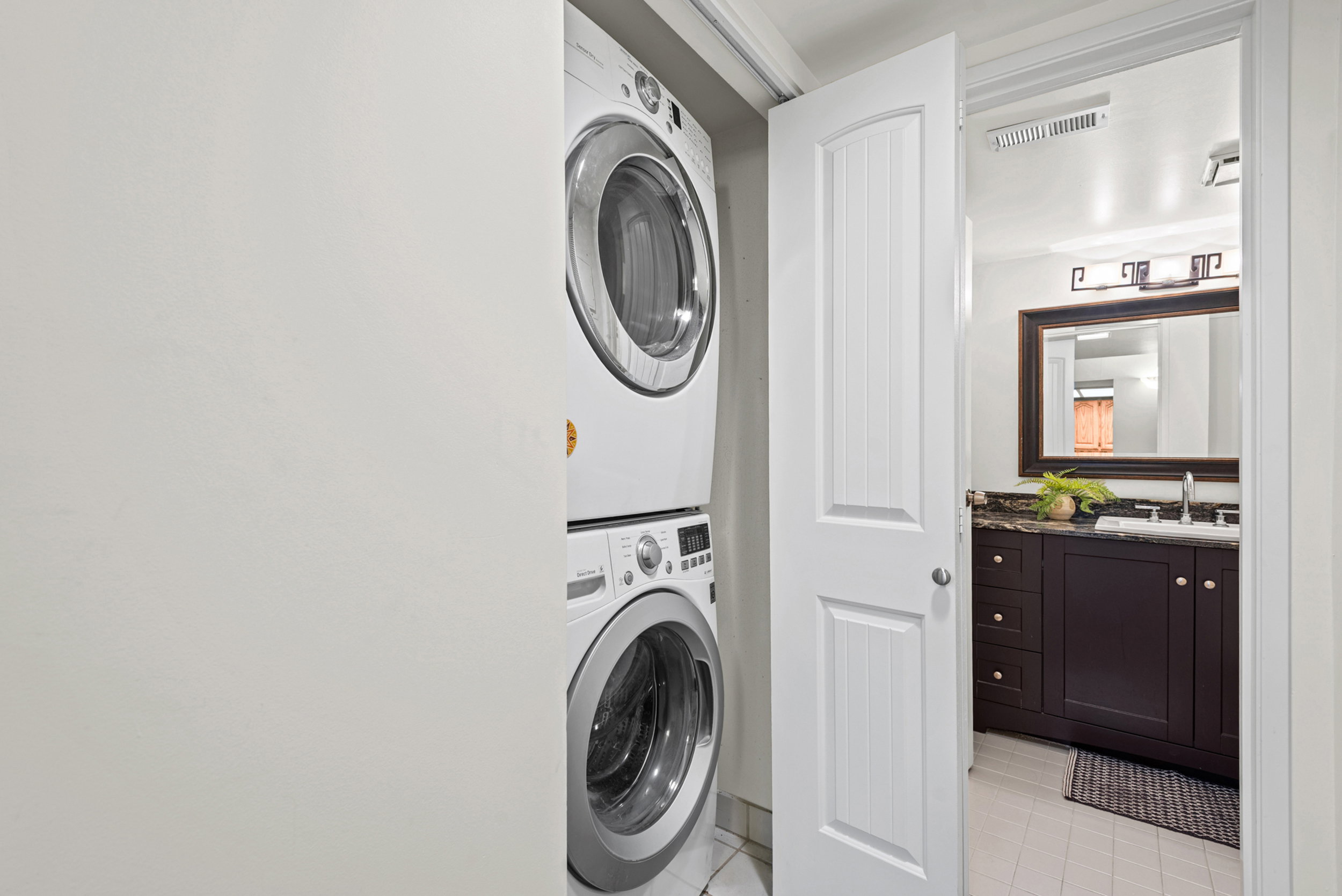 Stackable laundry appliances within a dedicated closet, with a paneled interior door opening to a bathroom featuring a dark wood-finish vanity, a stone countertop, an undermount sink, and an ornamental framed mirror