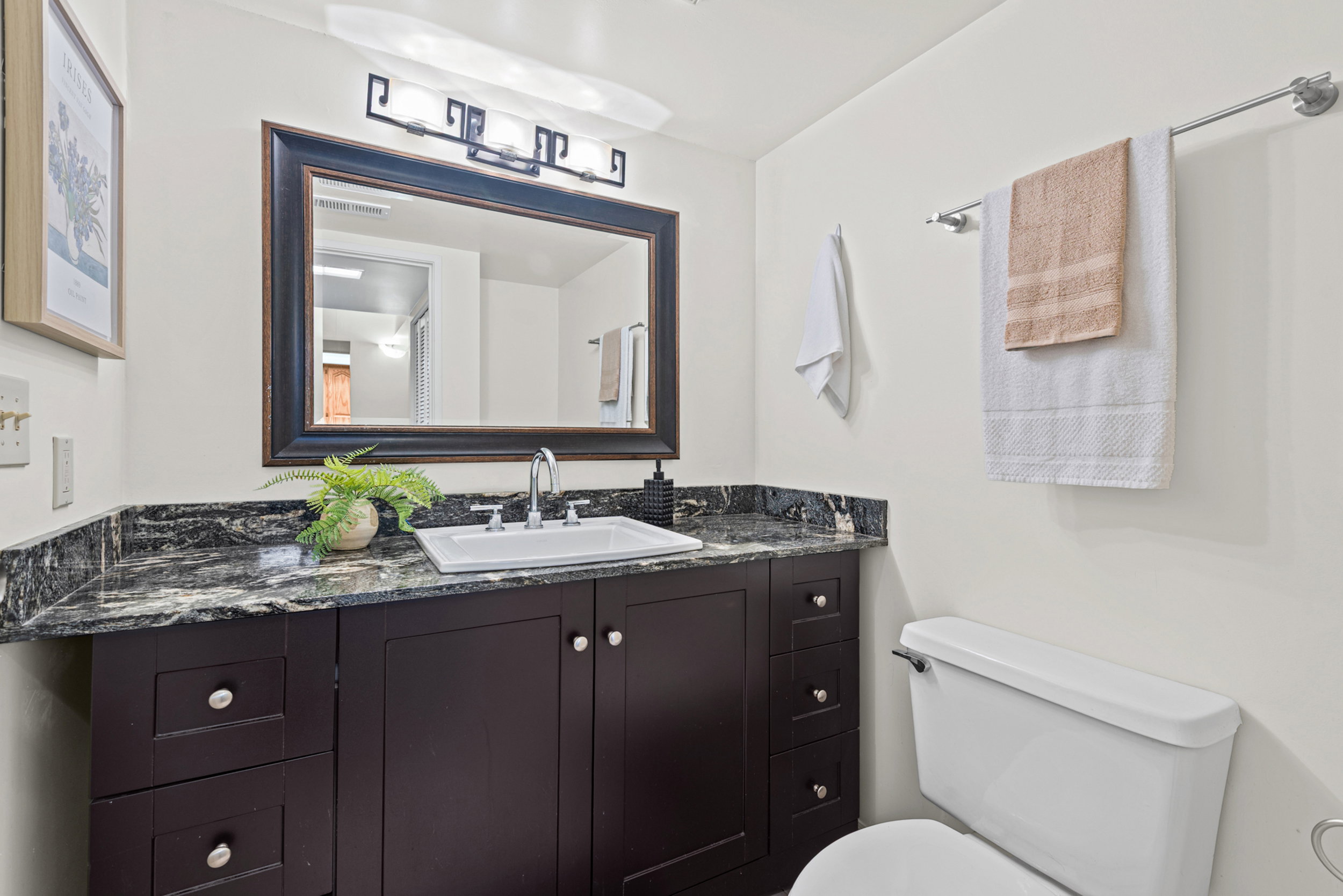 Dark wood-finish vanity with a stone countertop, an integrated rectangular sink, a framed mirror, and a three-light vanity fixture