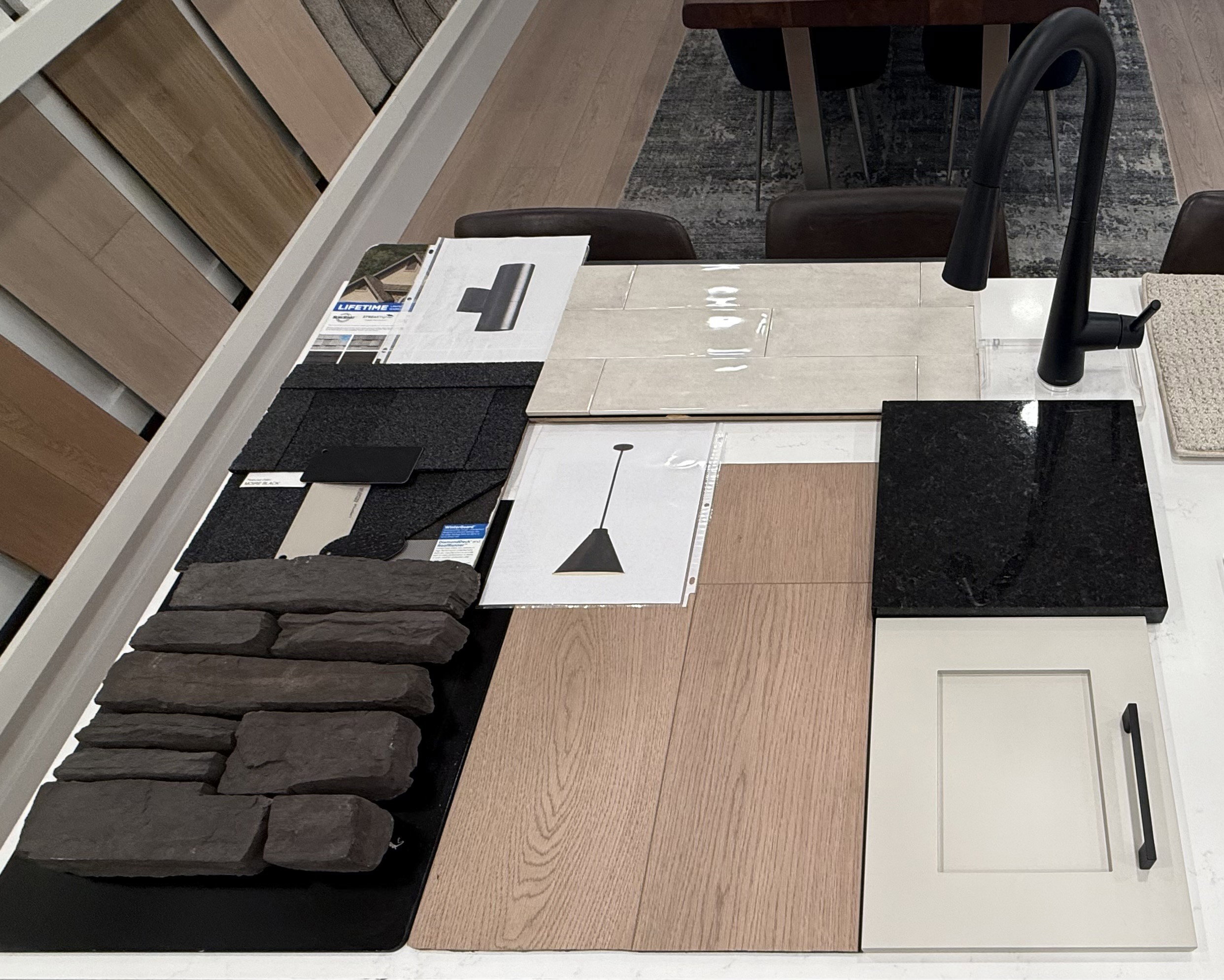 Selection of finishes including dark stone veneer, wood-finish flooring, black shingle roofing, glossy subway tile, and a cream shaker cabinet door with a matte black pull