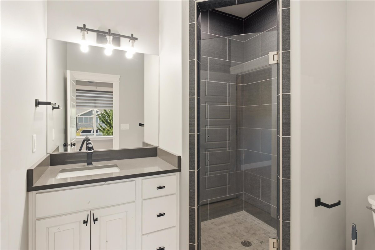 Bathroom featuring a glass-enclosed shower with dark tile surround, single vanity with light-toned countertop, white cabinetry with dark hardware, and a framed mirror with a horizontal light fixture