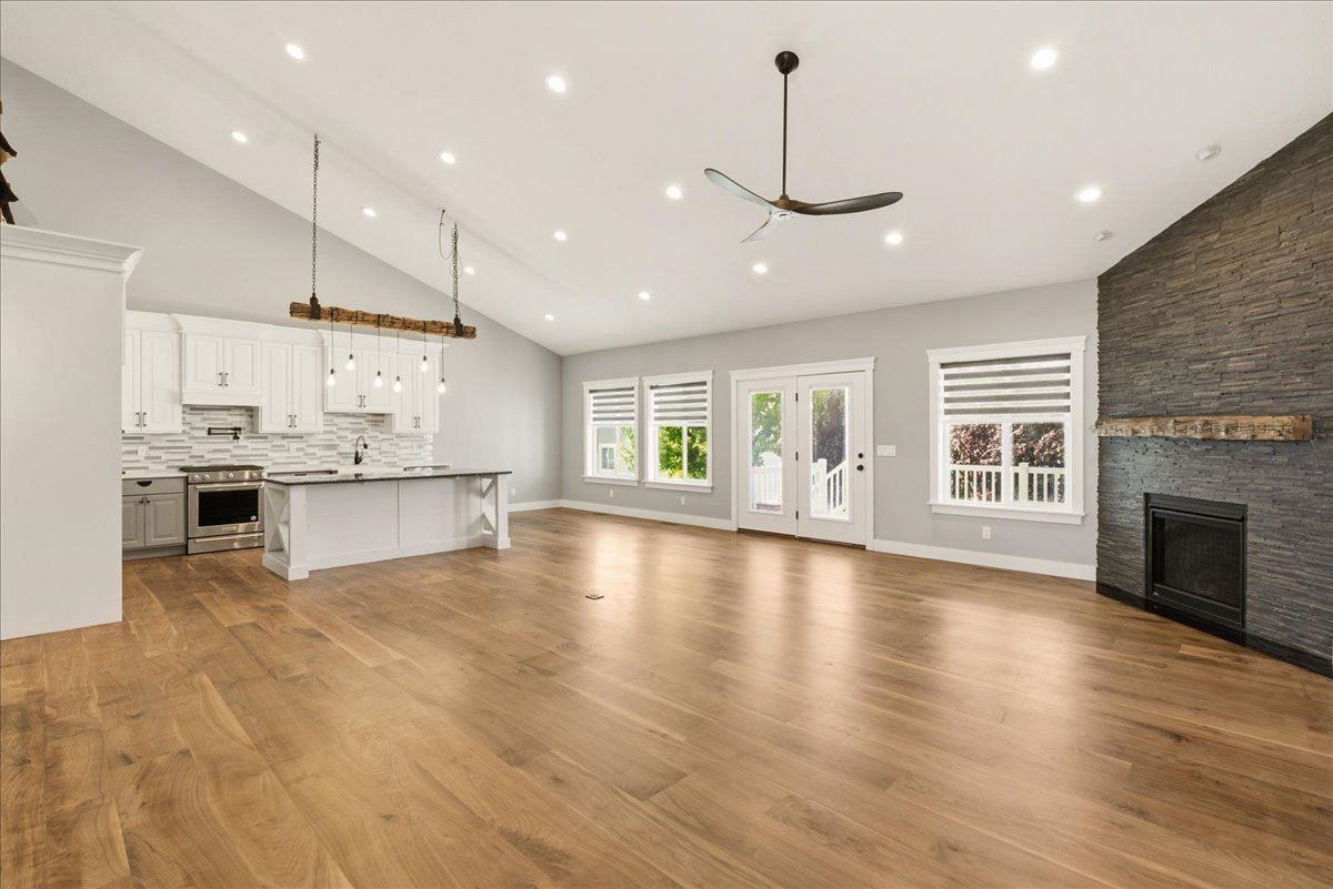 Open-concept living area featuring wood-finish flooring, a vaulted ceiling with recessed lighting and a ceiling fan, a stacked stone fireplace with a rustic mantel, and multiple windows with blinds