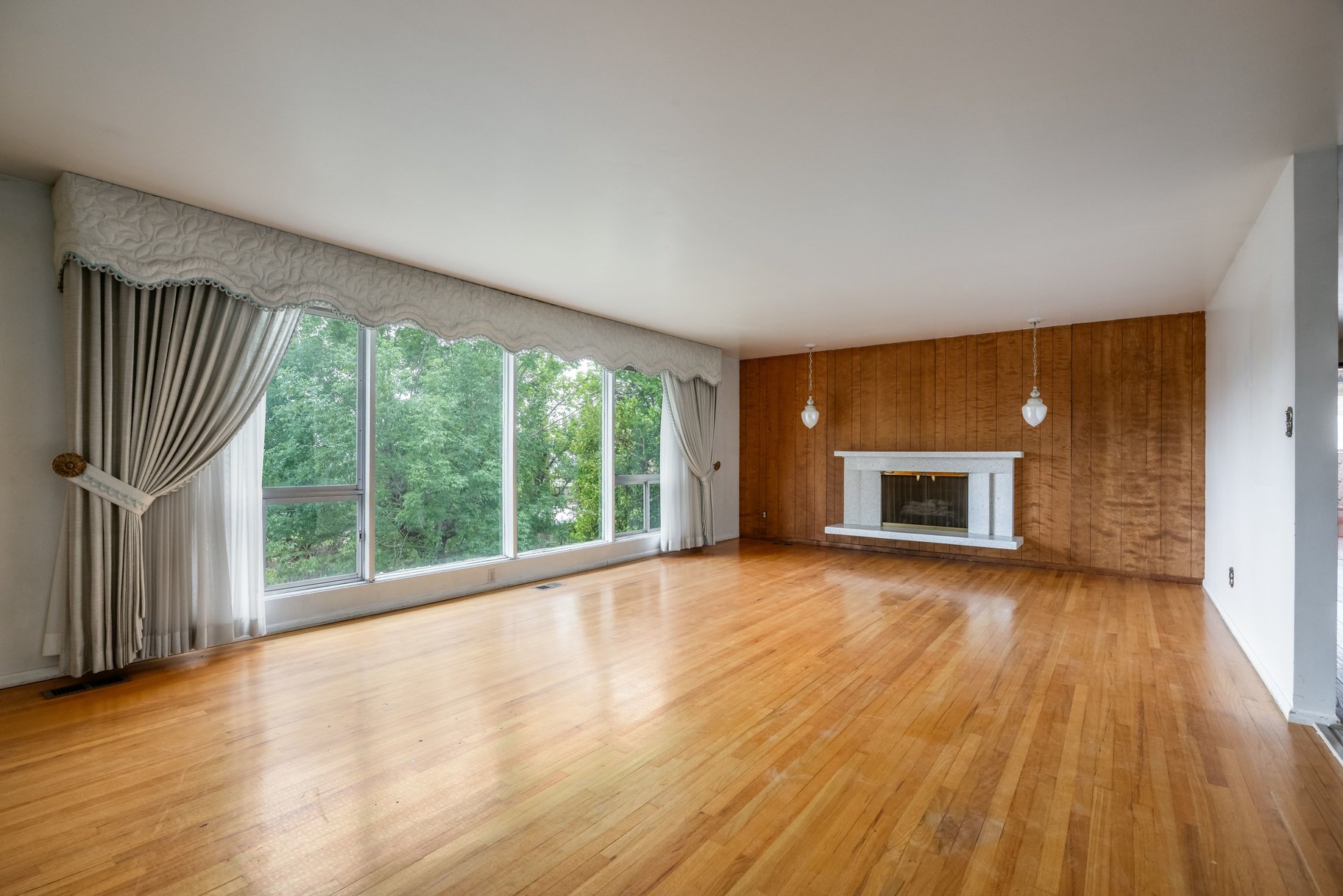 Spacious room featuring wood-finish flooring, a full wall of floor-to-ceiling windows, and a white mantel fireplace with a wood panel accent wall
