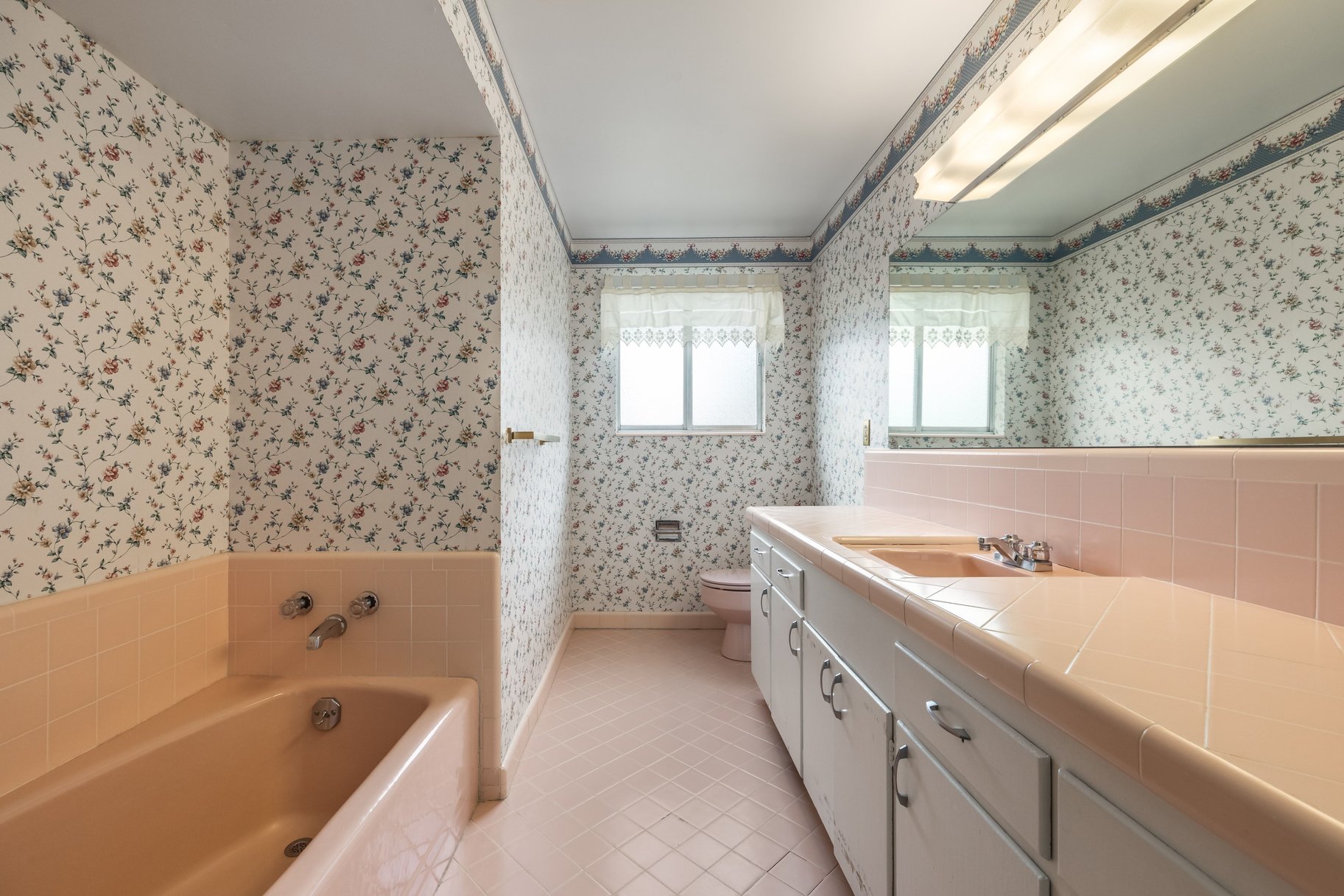 Bathroom featuring a built-in tub, a vanity with dual windows, a large mirror, and patterned wallpaper