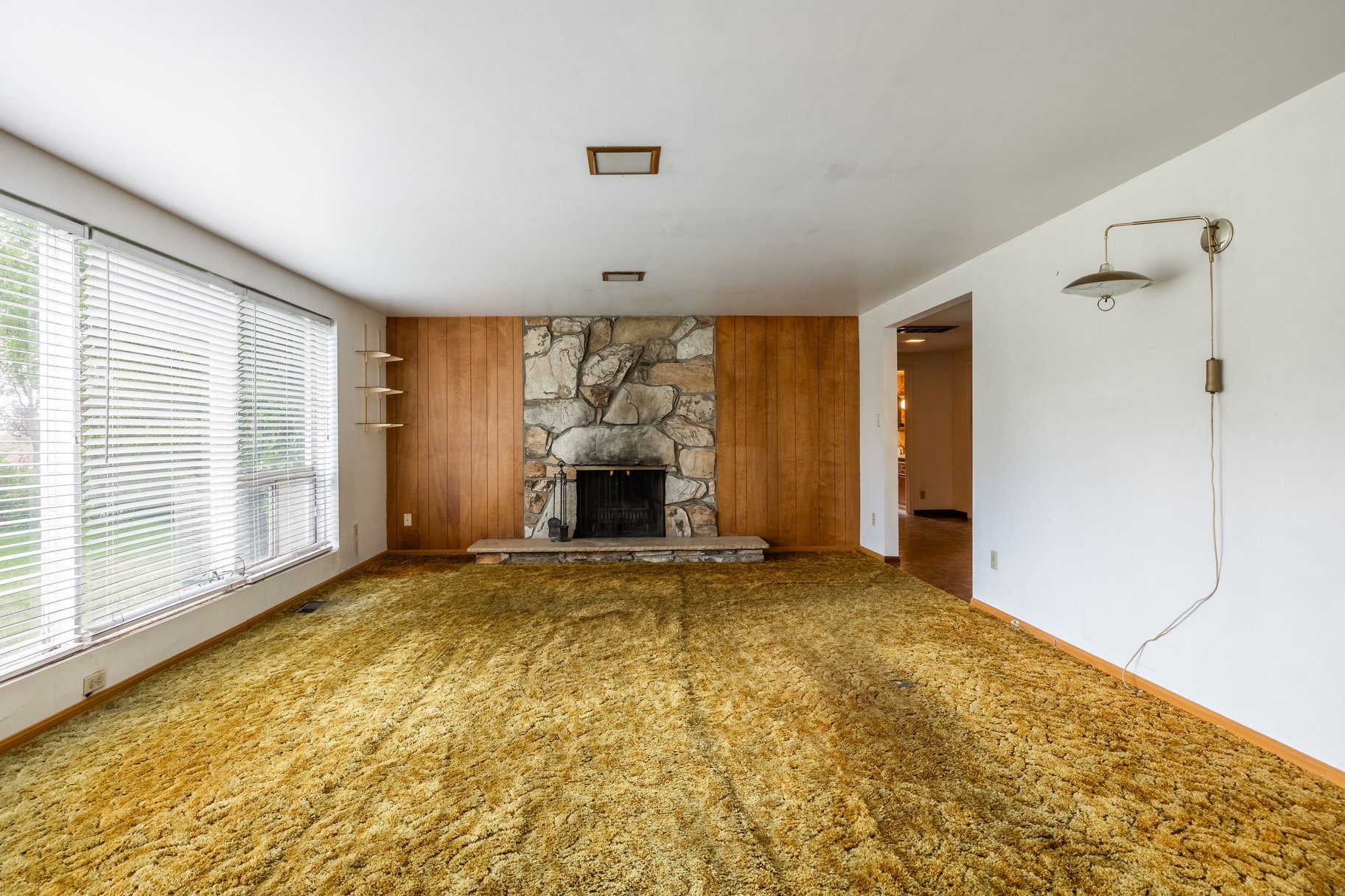 Living room featuring a stone fireplace with a raised hearth, wood paneling, and a large window with blinds
