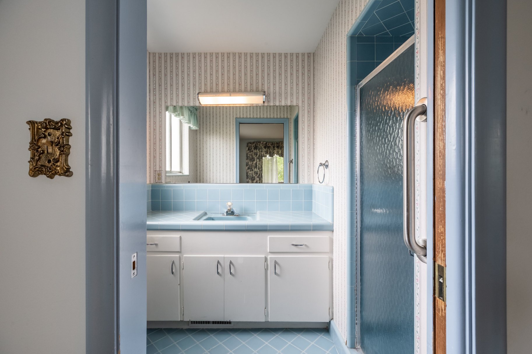 Bathroom featuring blue ceramic floor tiles, a white vanity with blue countertop and sink, light-toned striped wallpaper, and a shower enclosure with a textured glass door