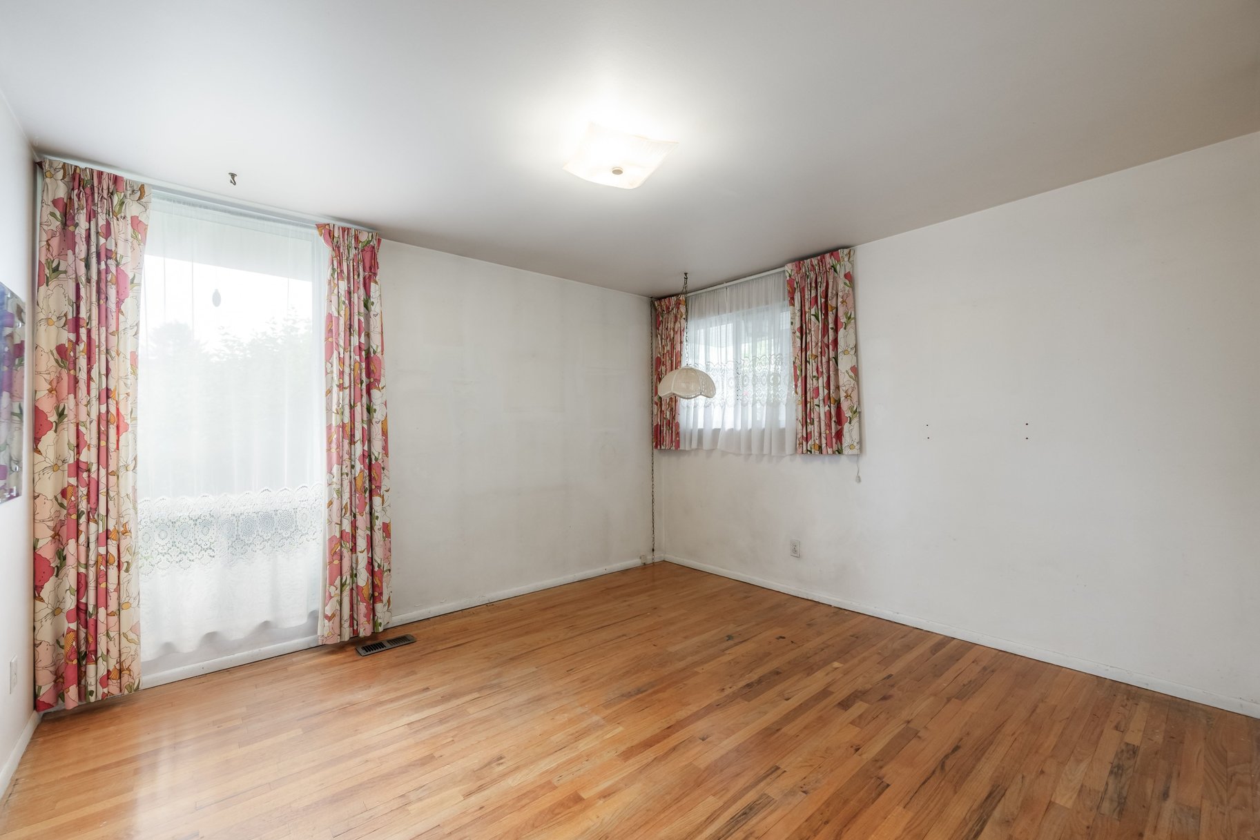 Expansive room featuring wood-finish flooring, white walls, and a large window with sheer and floral curtains