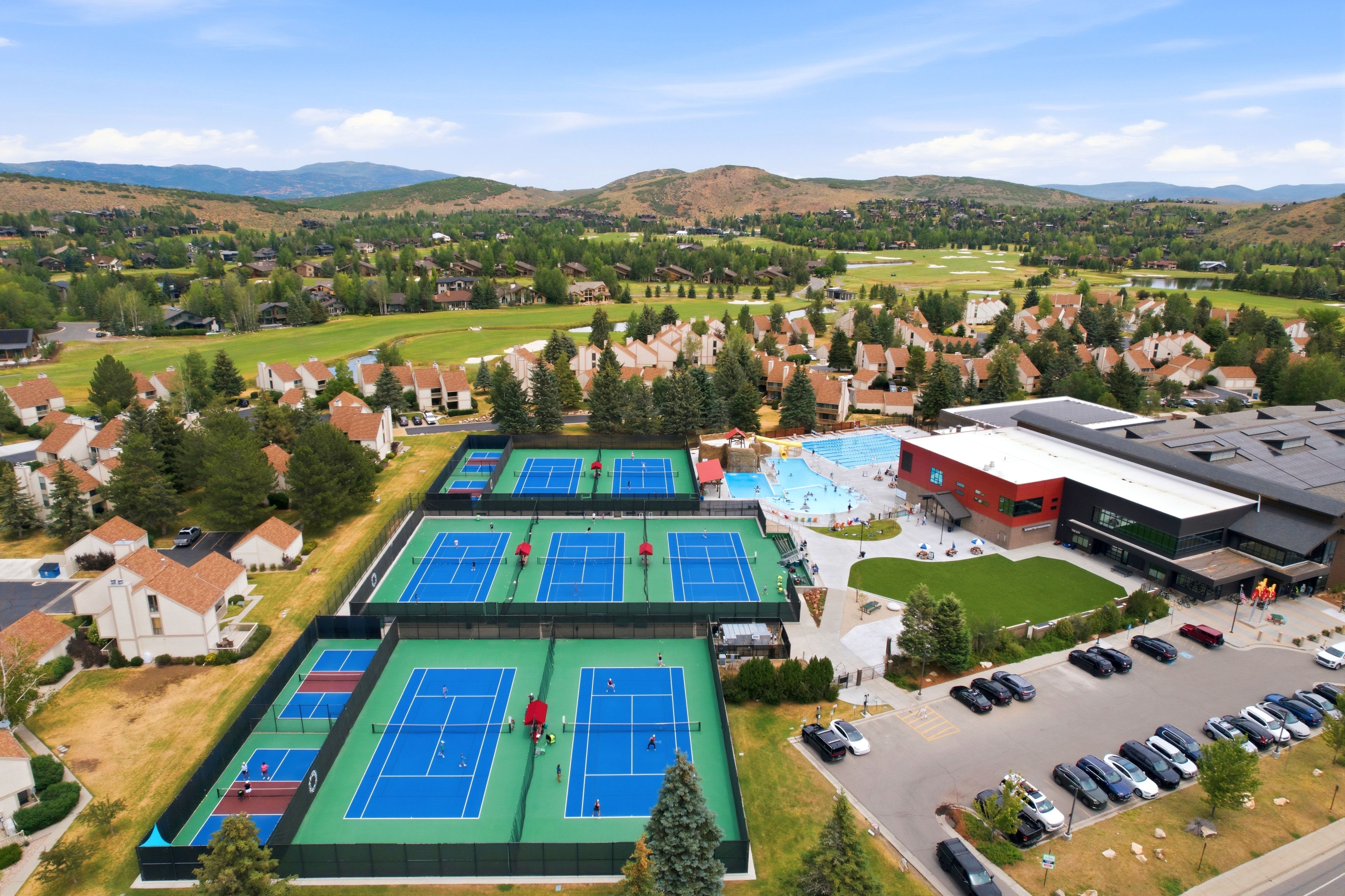 Outdoor recreational complex featuring multiple tennis courts, a large swimming pool area, expansive green spaces, and a substantial recreation center building with ample parking
