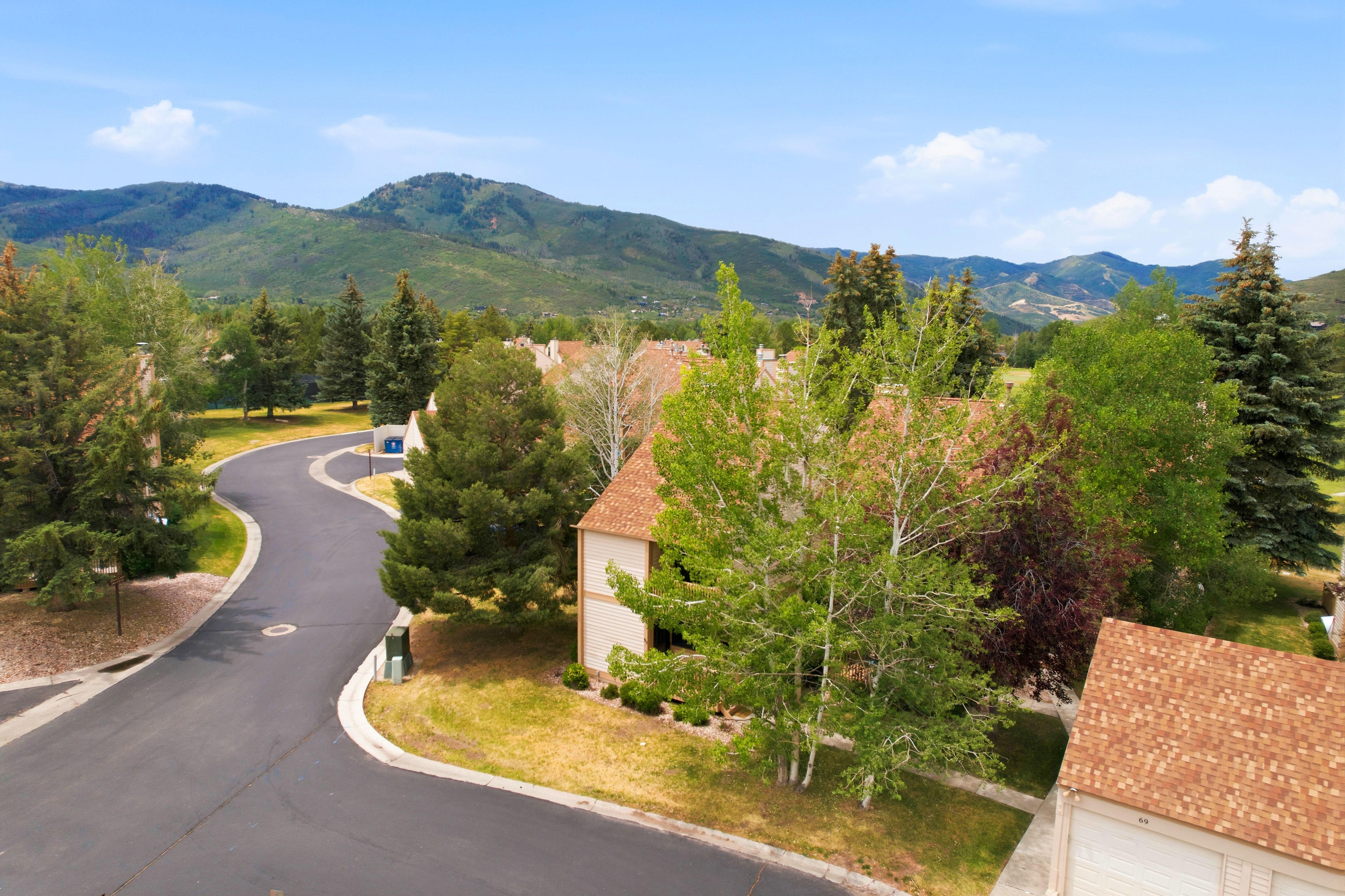 Mountainous landscape framing the property, featuring a paved road with curbs, established trees, and residential structures with shingle roofs