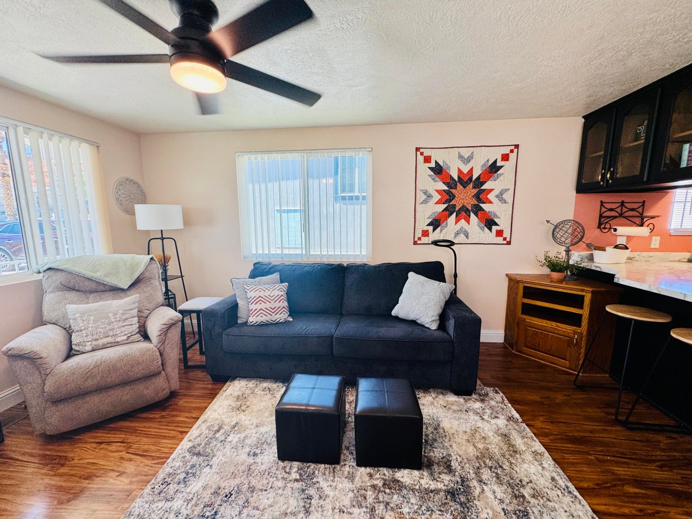 Living area featuring wood-finish flooring, a ceiling fan with integrated lighting, and multiple windows with vertical blinds