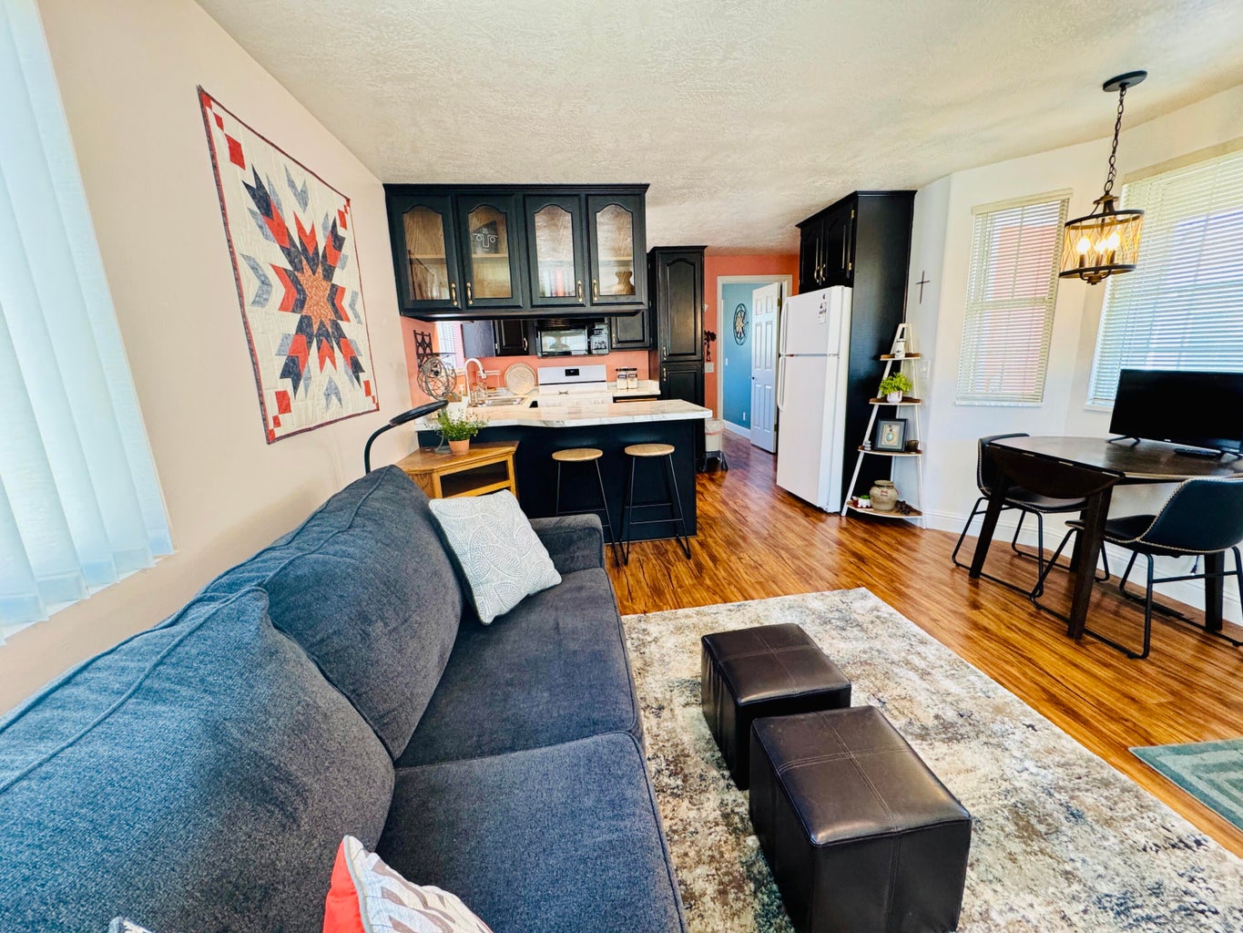 Open-concept living area featuring rich wood-finish flooring, a kitchen with dark cabinetry and a light-colored countertop, and a dining area illuminated by a decorative pendant light