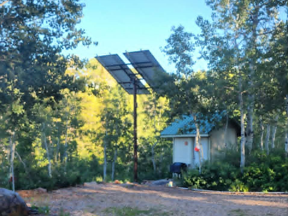 Gravel area featuring a standalone solar panel array and a small outbuilding with a green roof, surrounded by mature trees
