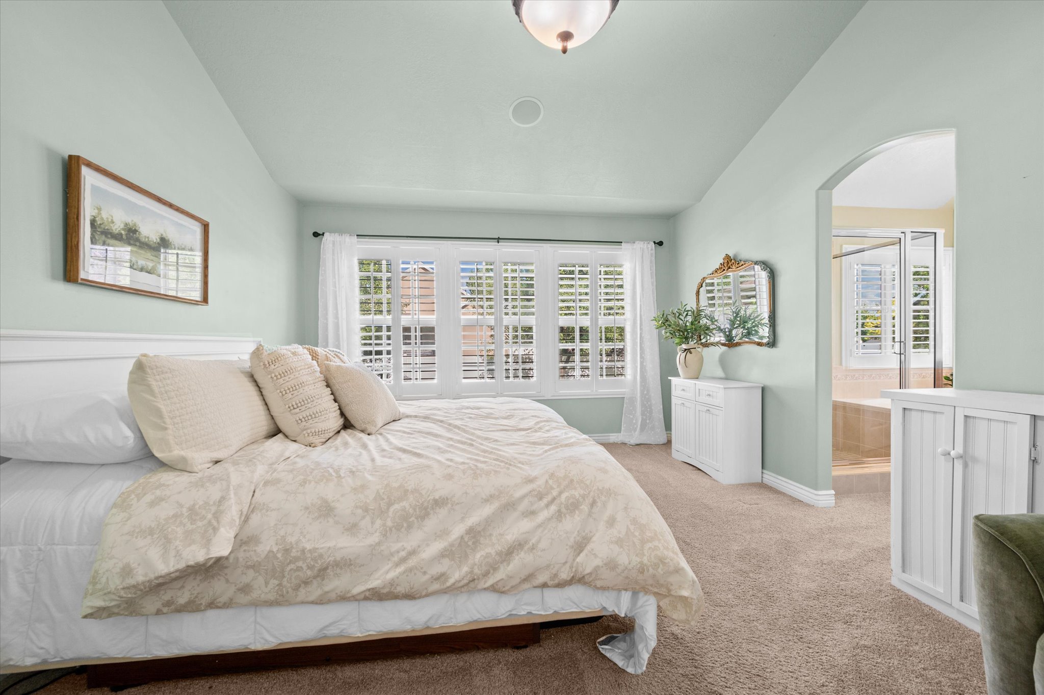 Vaulted ceiling bedroom featuring plantation shutters, carpet flooring, and a wide arched entryway revealing a bathroom with a tiled surround tub