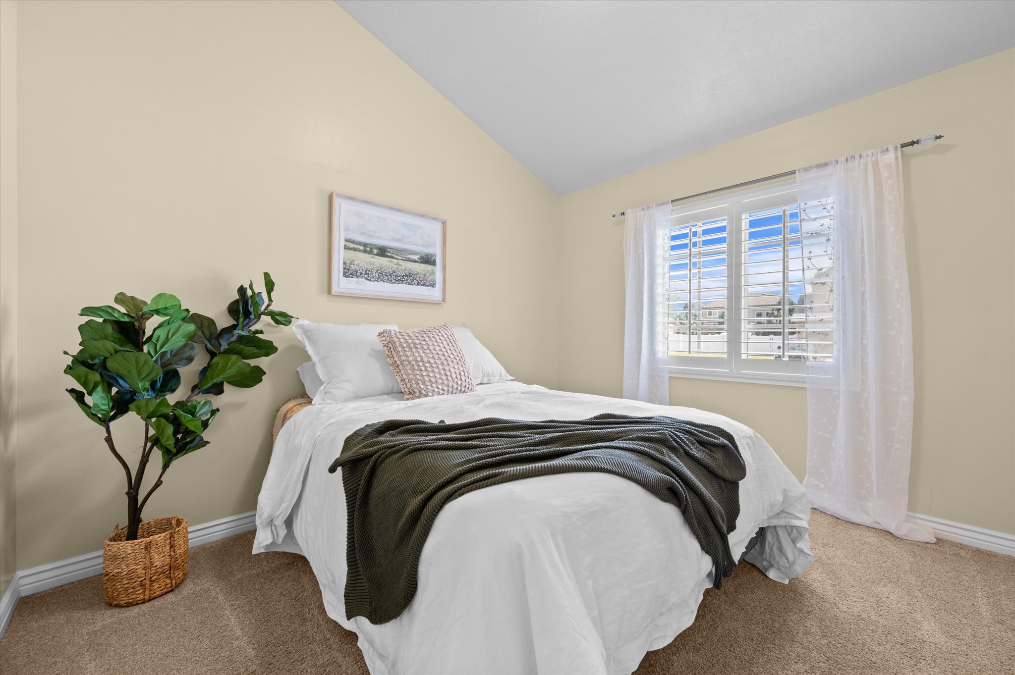 Vaulted ceiling bedroom with a single window featuring plantation shutters and sheer white curtains