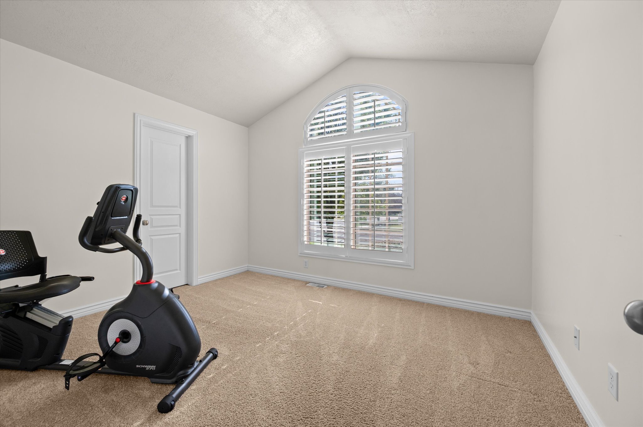 Vaulted ceiling room featuring a large arched window with plantation shutters, light-colored carpet flooring, white baseboards, and a paneled interior door