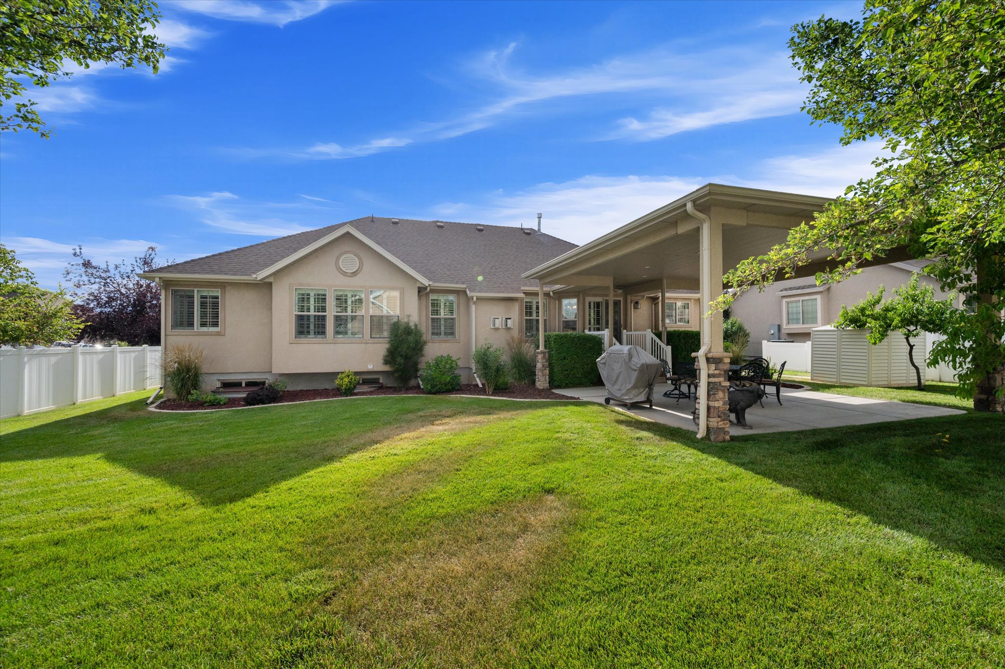 Expansive backyard featuring a covered patio, concrete hardscaping, mature trees, manicured lawn, and a white privacy fence