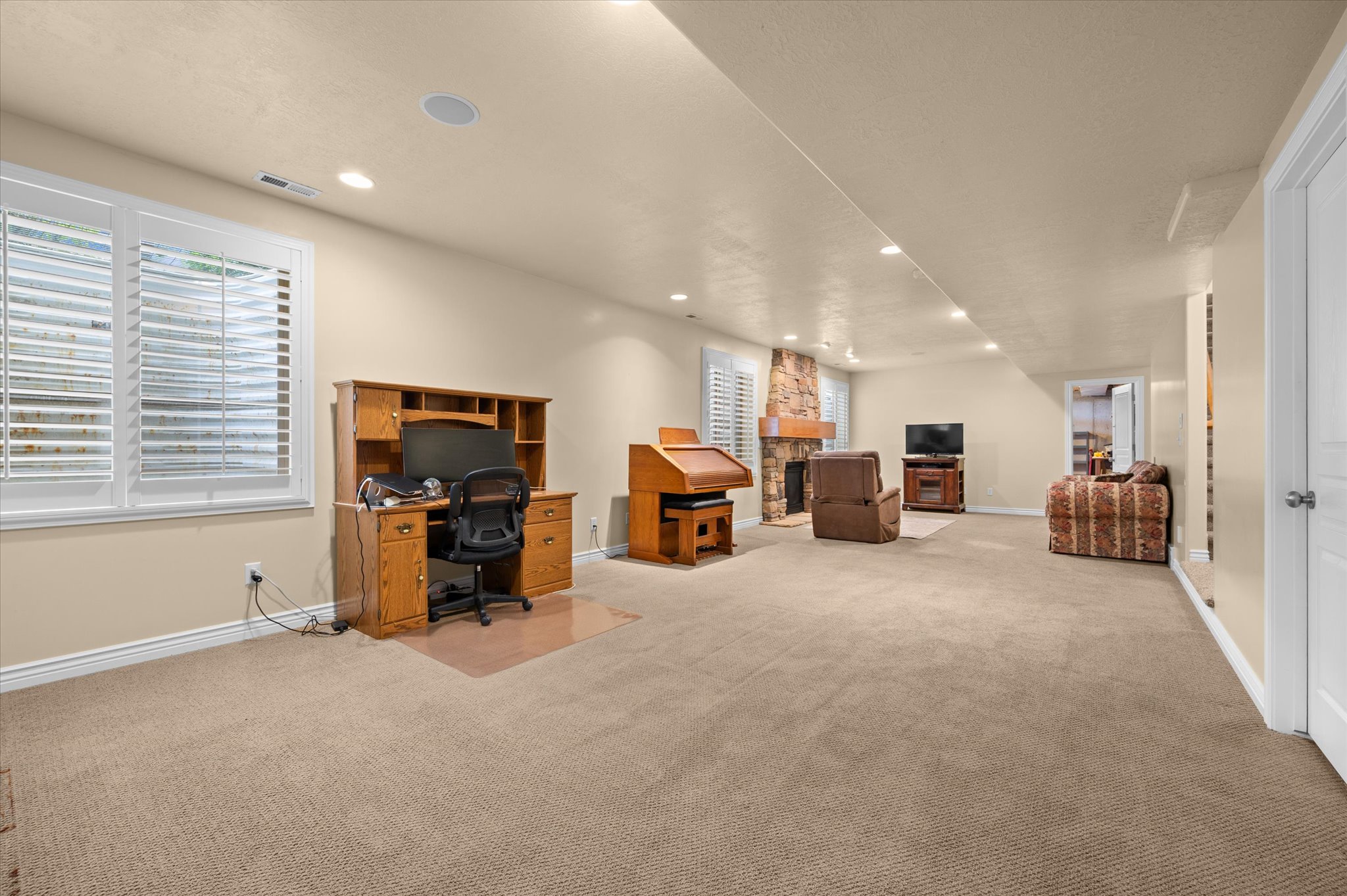 Spacious carpeted room featuring recessed lighting, a stone fireplace, and multiple windows with plantation shutters