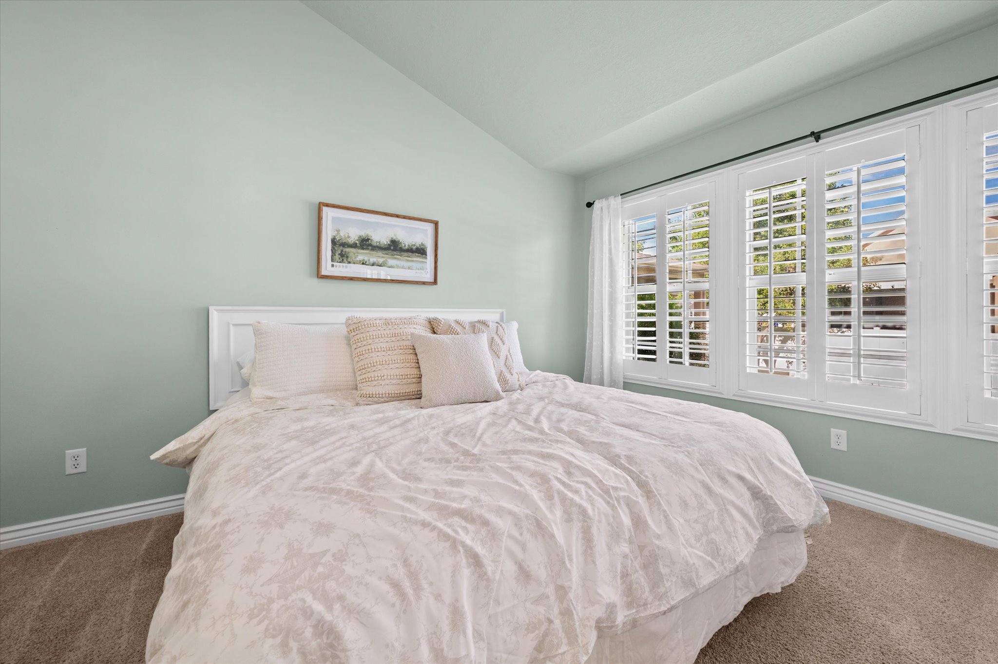 Vaulted ceiling bedroom featuring plantation shutters, light green walls, white trim, and neutral-toned carpeting