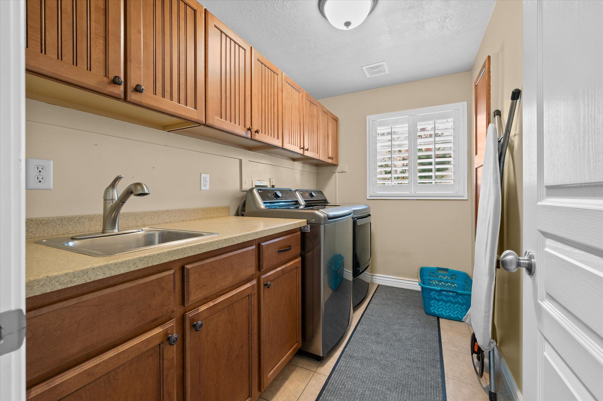 Dedicated utility space featuring extensive wood cabinetry, a practical sink with a chrome faucet, light-toned countertop, and a window with white louvered shutters