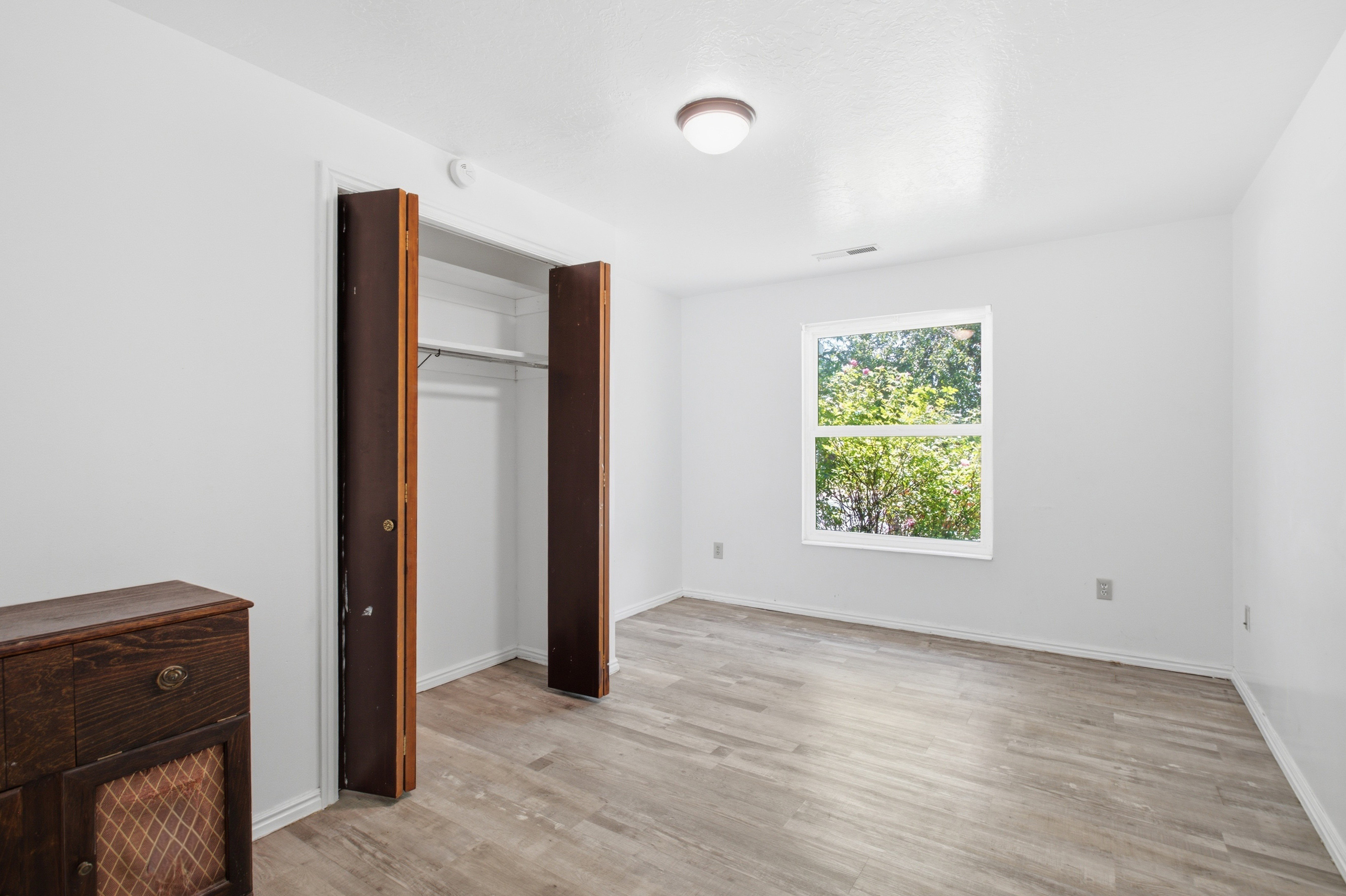 Room featuring neutral wall paint, wood-finish flooring, a window providing natural light, a ceiling-mounted light fixture, and an open closet with a hanging rod