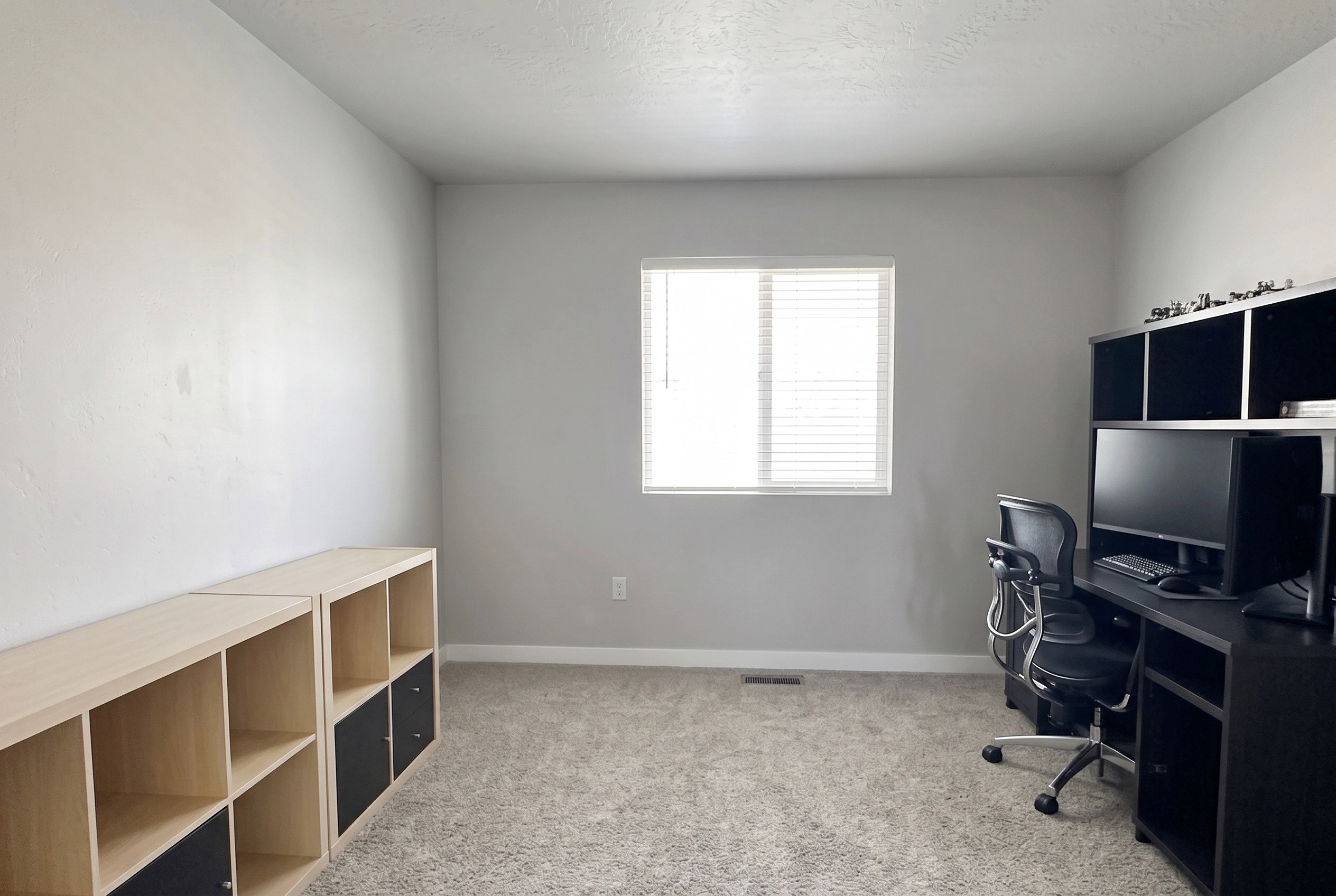 Neutral-toned interior room featuring light gray walls, textured carpet flooring, and a window with horizontal blinds