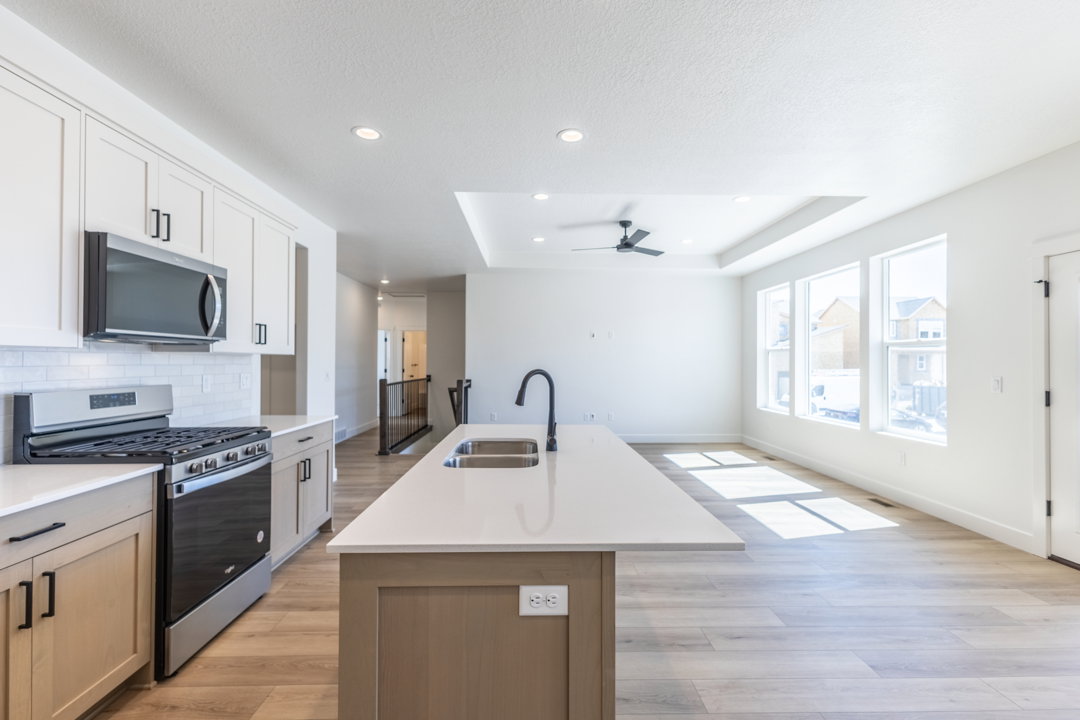 Open-concept kitchen and living area featuring a large center island with double sink, stainless steel appliances, light wood-finish flooring, white cabinetry, and a tray ceiling with a ceiling fan