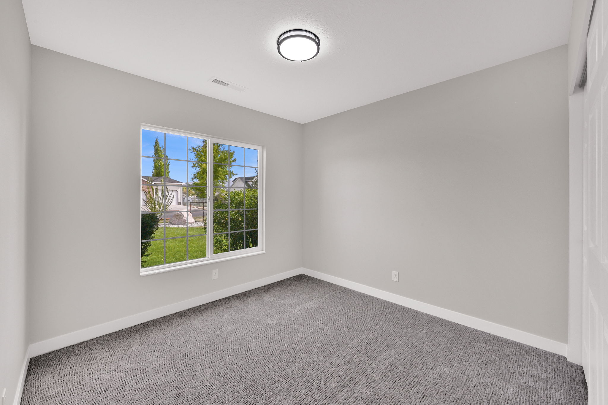 Bright room featuring light gray walls, a white-framed window with grid panes, textured gray carpeting, white baseboards, and a contemporary flush-mount ceiling light fixture