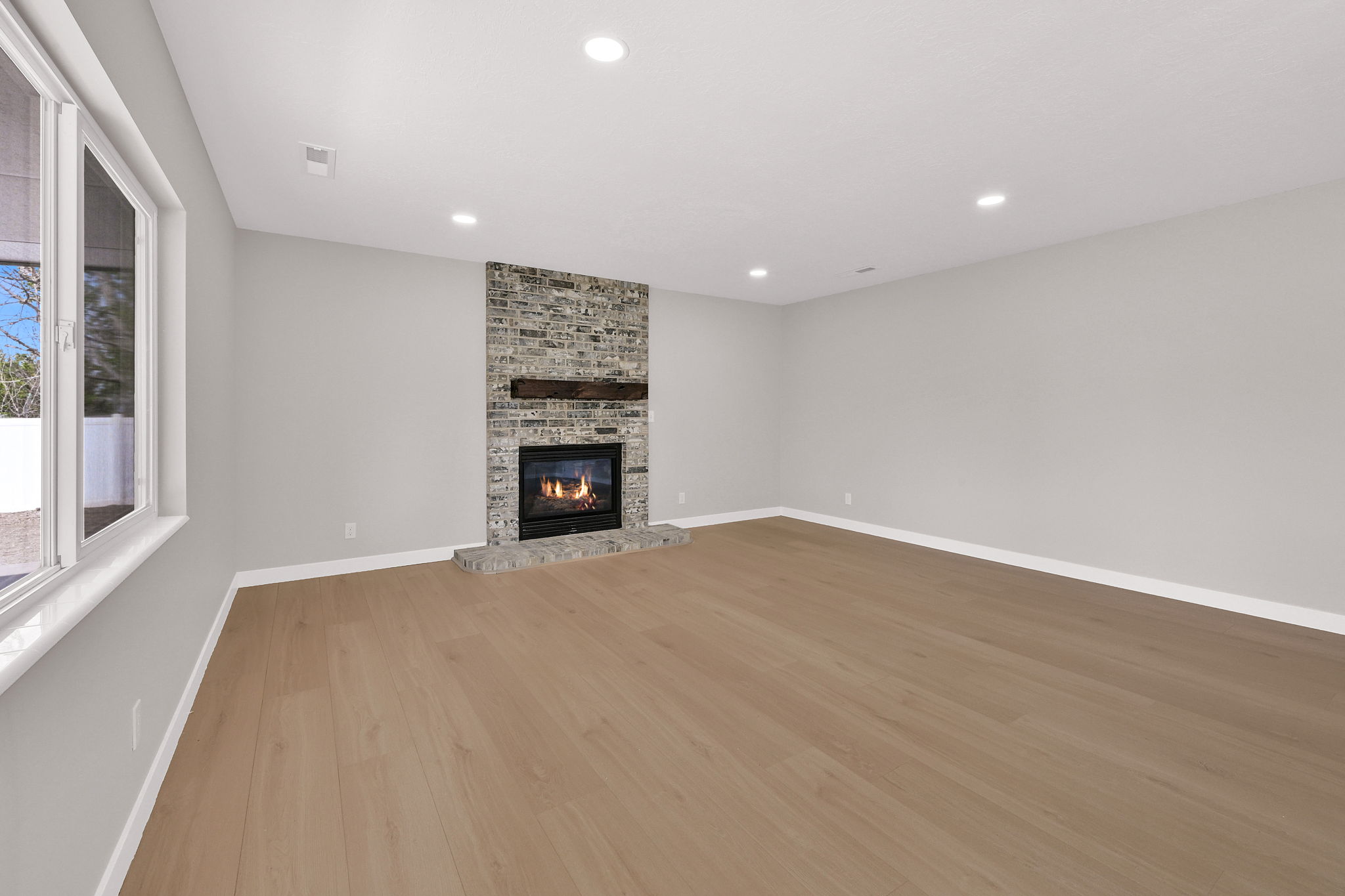 Spacious room featuring light wood-finish flooring, a stone-clad fireplace with a dark wood mantel, recessed lighting, and large windows