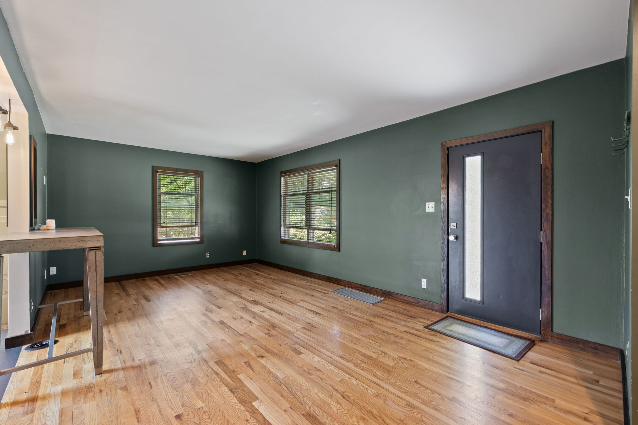 Spacious room featuring wood-finish flooring, two large windows with blinds, a built-in counter, and a modern entry door with a vertical glass insert