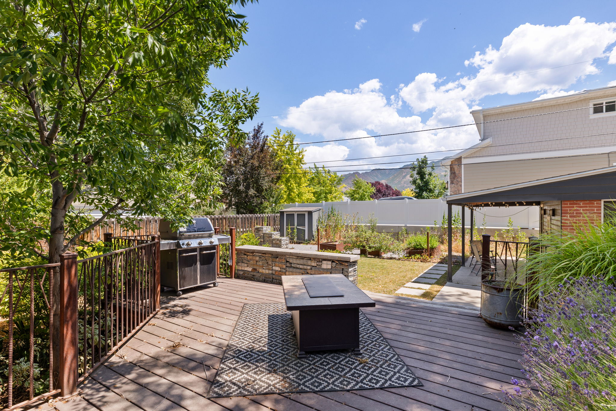 Expansive outdoor living area featuring a spacious wood-finish deck, stone retaining walls, mature landscaping, and mountain scenery