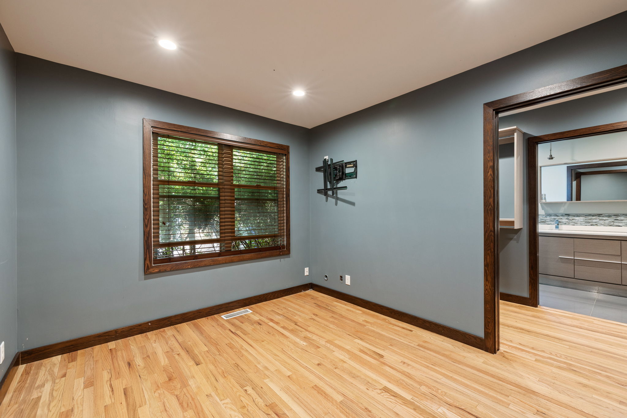 Room featuring hardwood flooring, recessed lighting, a window with wood blinds, and a doorway opening to a bathroom with a vanity