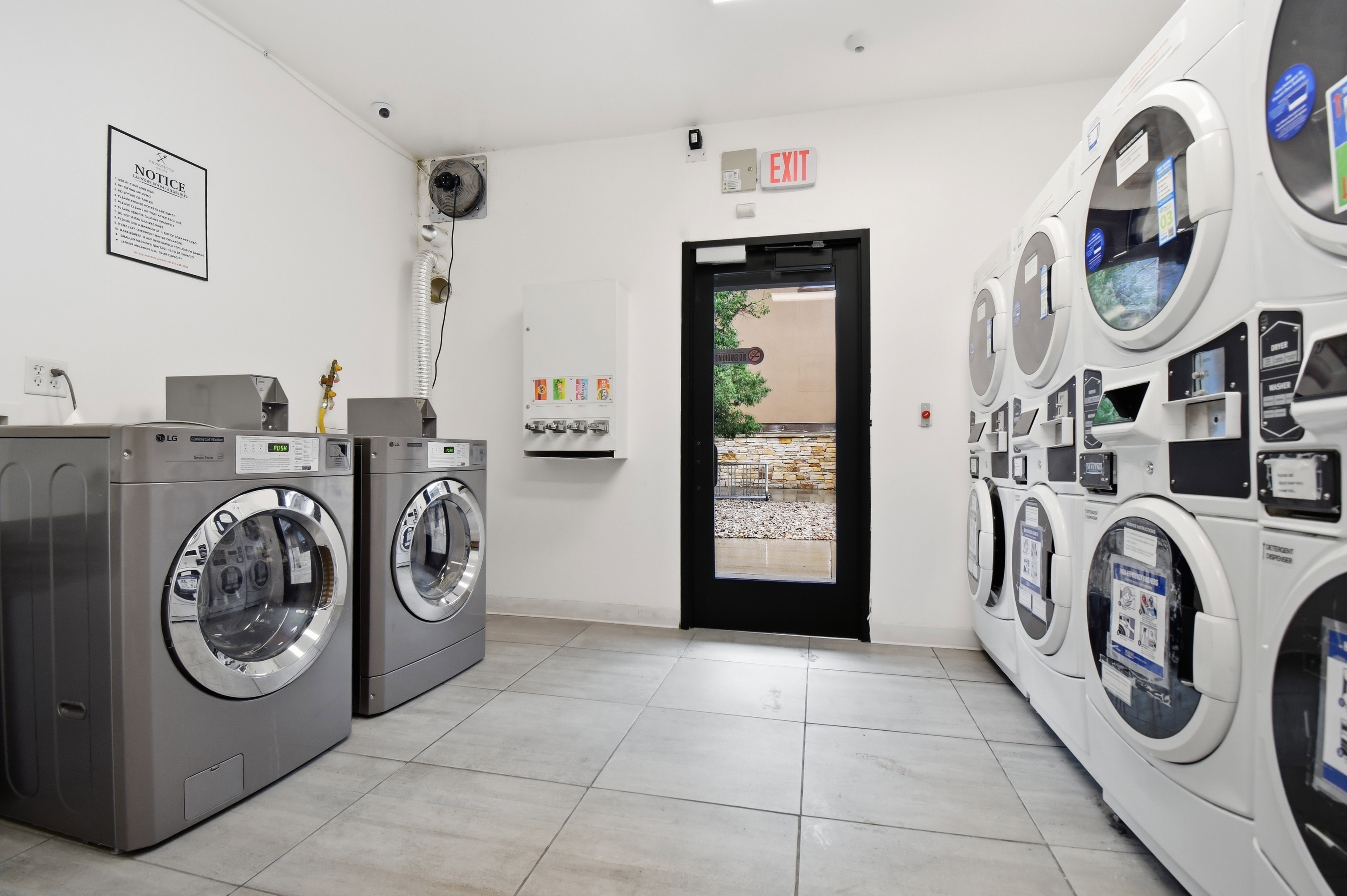 Dedicated laundry room featuring multiple front-loading washers and dryers, light-colored tile flooring, white walls, and a black-framed glass exit door