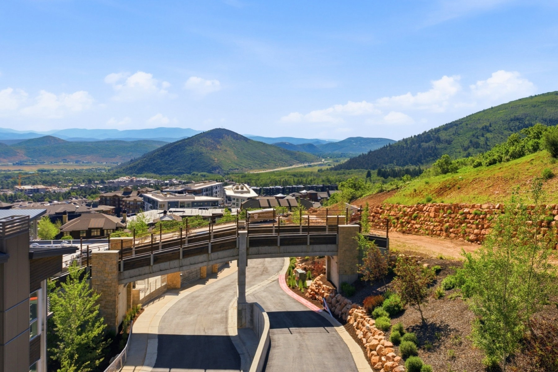 Elevated pedestrian bridge over a paved roadway, surrounded by tiered landscaping with natural stone retaining walls and mature greenery