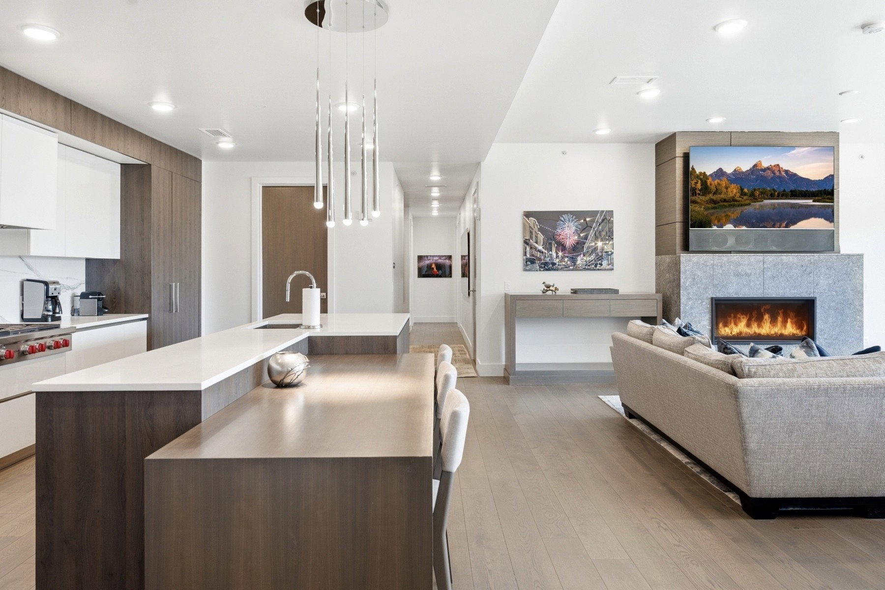 Open-concept living space with wood-finish flooring, a contemporary kitchen island featuring a waterfall countertop, and a built-in linear fireplace with a stone surround