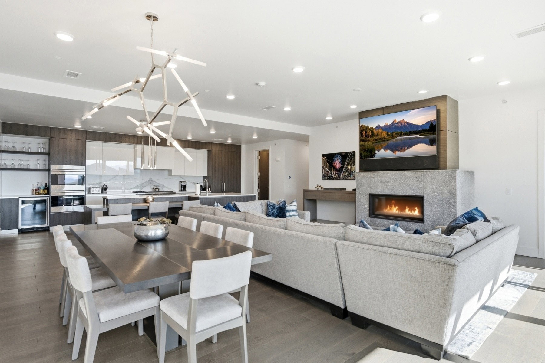 Open-concept living area featuring wood-finish flooring, a contemporary linear fireplace with a stone surround, a modern chandelier, recessed lighting, and a kitchen with integrated appliances and a beverage cooler