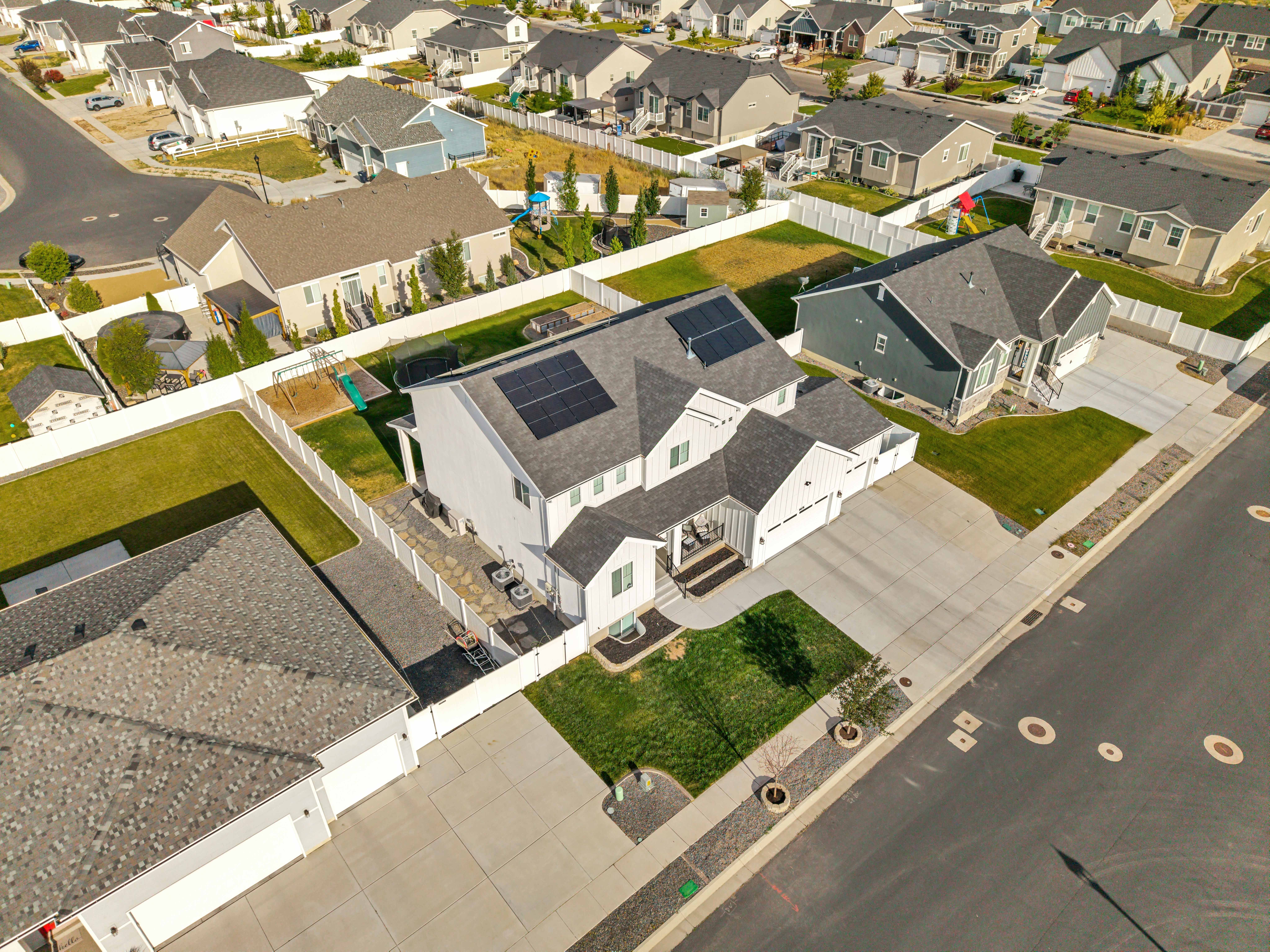 Contemporary white siding residence featuring a dark shingle roof with integrated solar panels, an attached garage, a spacious concrete driveway, and a fenced backyard with a patio
