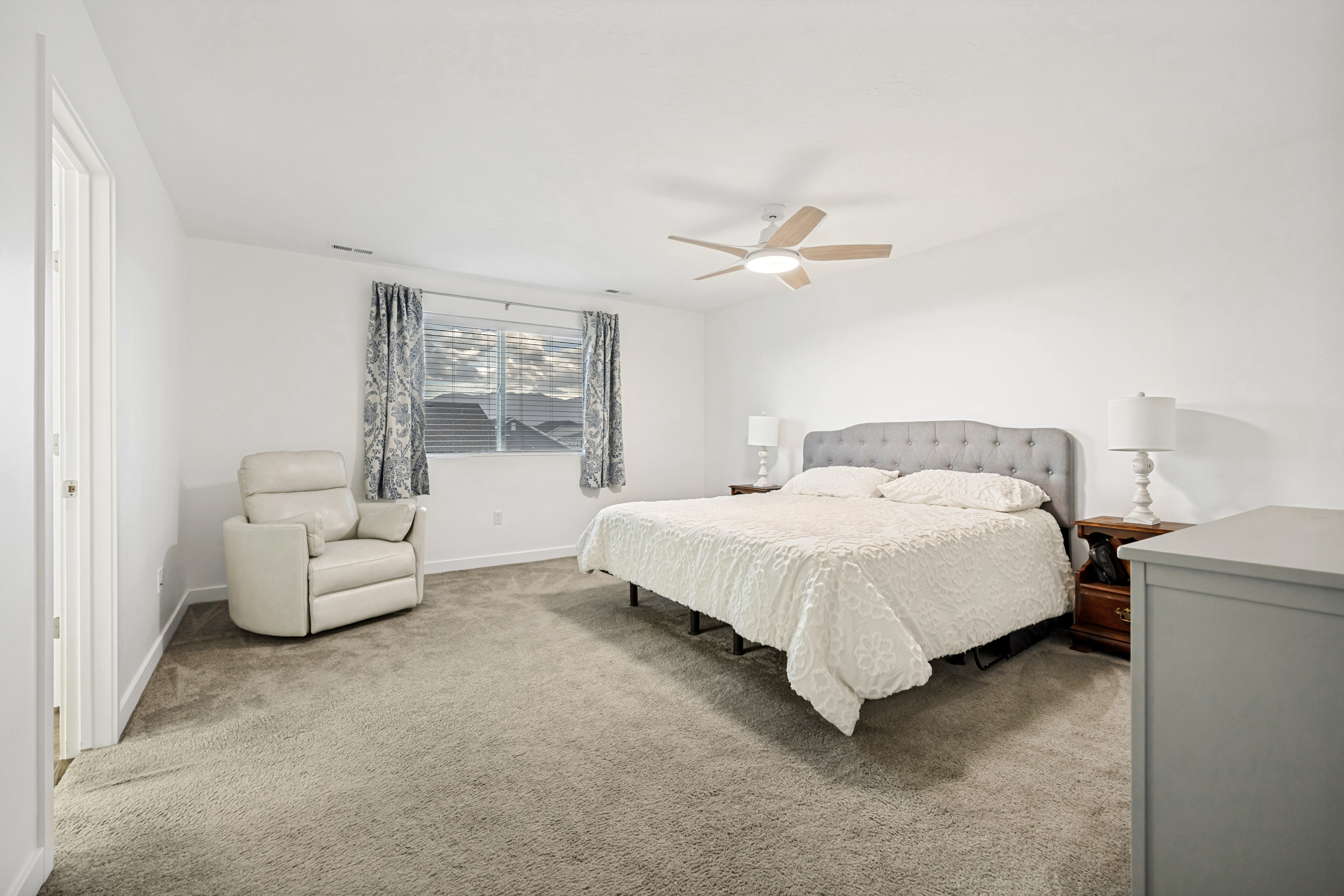 Carpeted bedroom with a ceiling fan light fixture, two windows with blinds and patterned curtains, and a light gray tufted headboard
