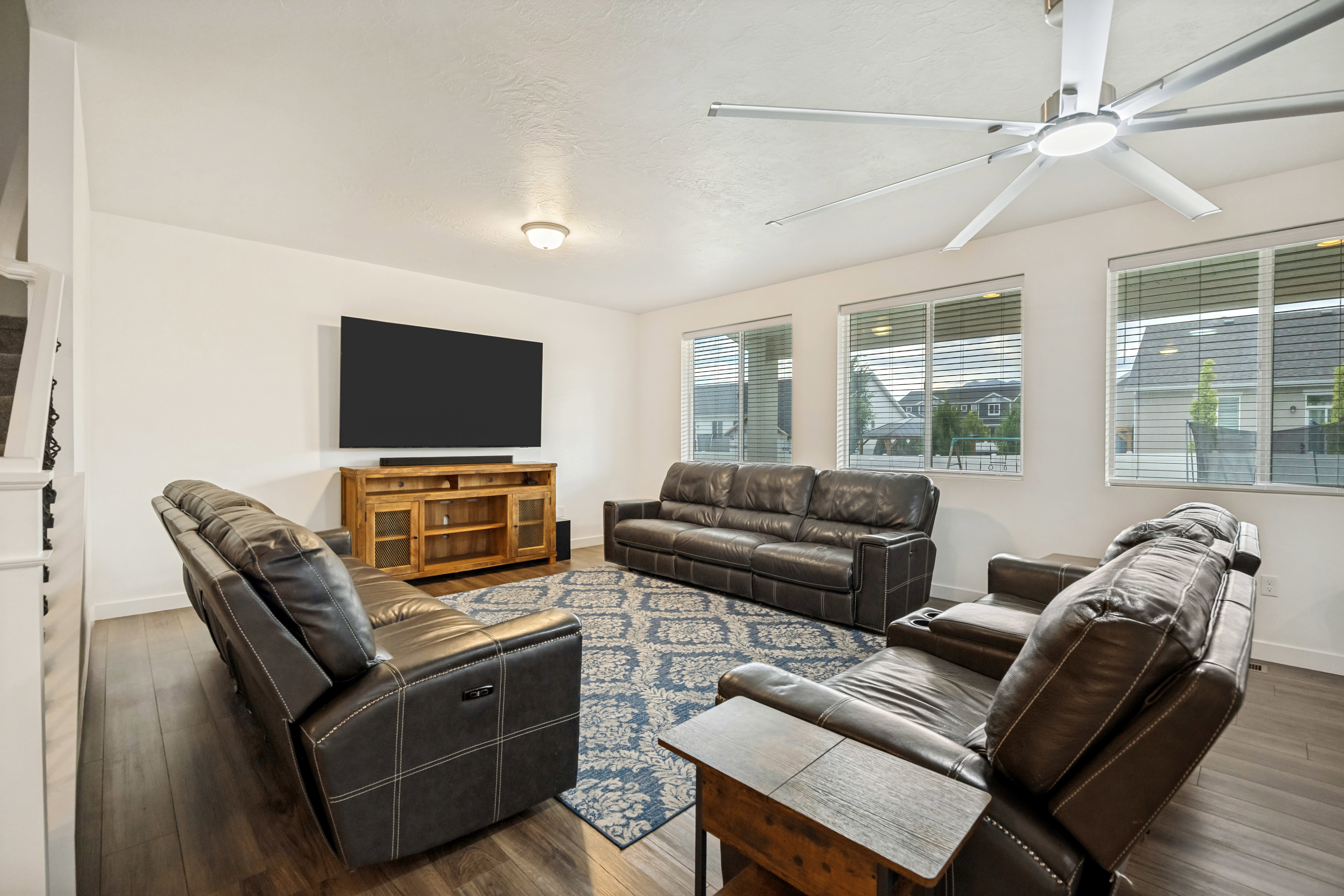 Spacious entertainment room featuring wood-finish flooring, a contemporary ceiling fan, and multiple windows with blinds
