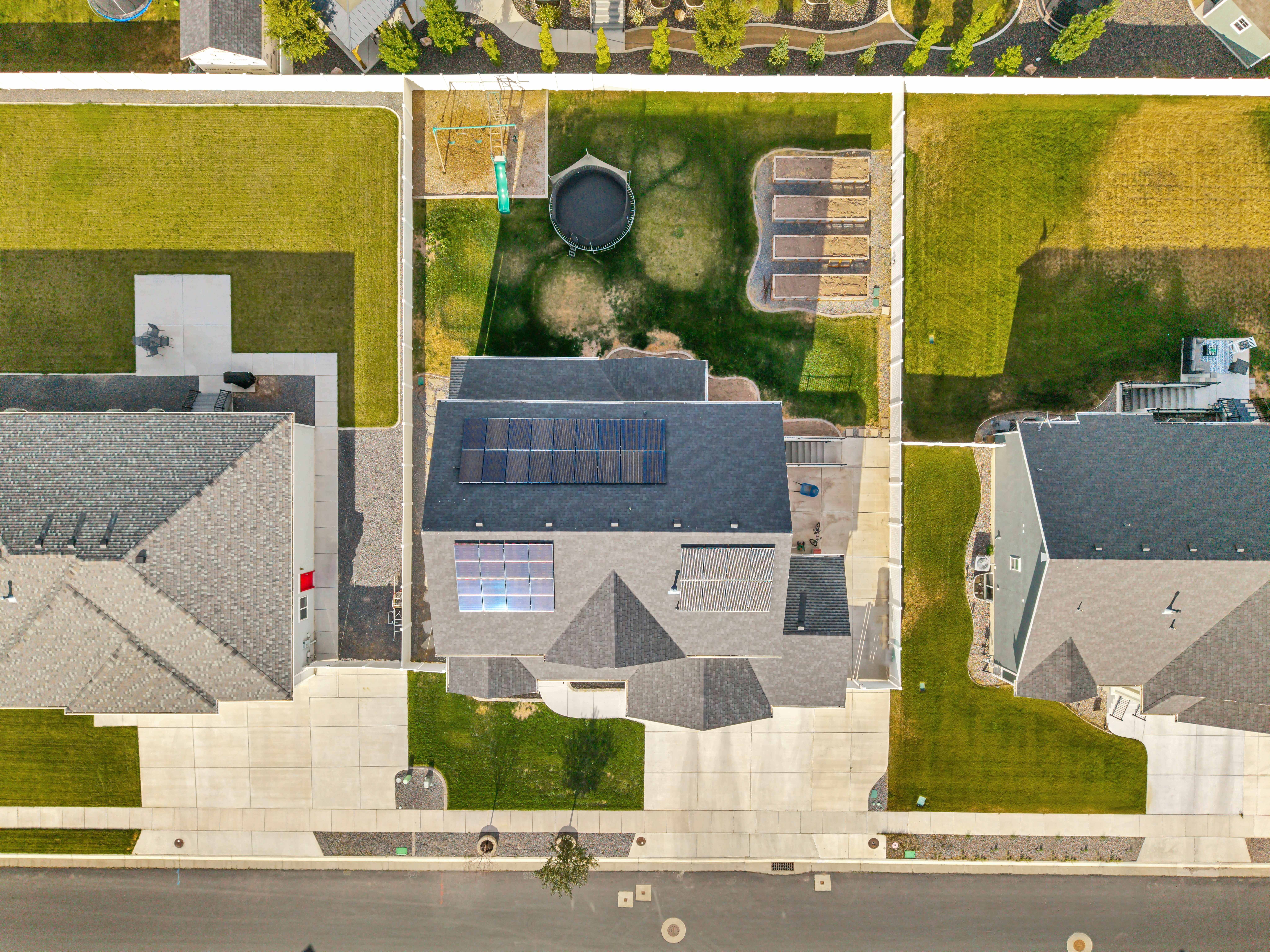 Aerial view of a residential property featuring solar panels on the roof, a fenced backyard with raised garden beds, and a concrete patio