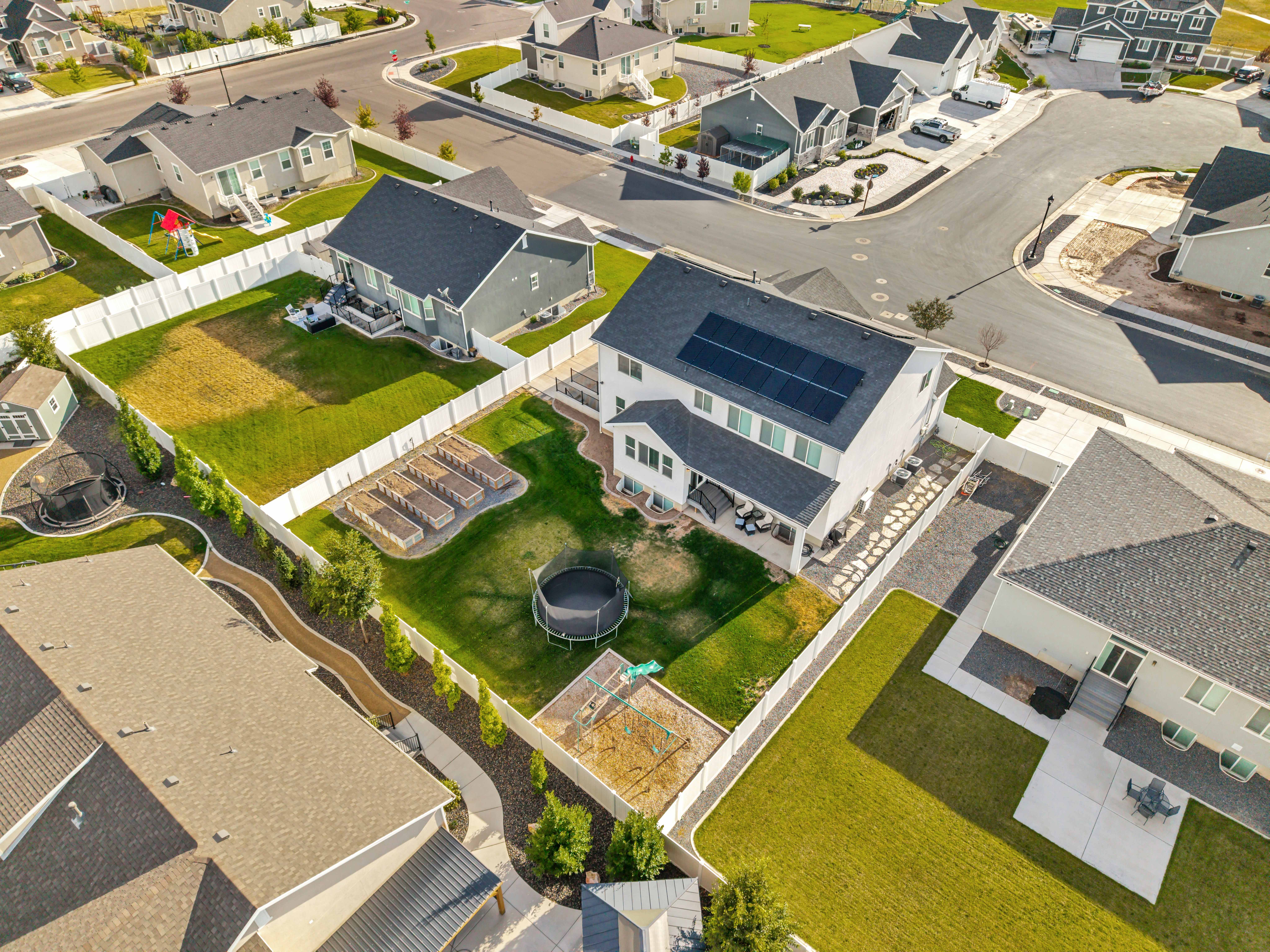Aerial perspective of a contemporary residence featuring solar panels, an expansive fenced yard, an established garden area with raised beds, a stone-paved patio, and a dedicated play area