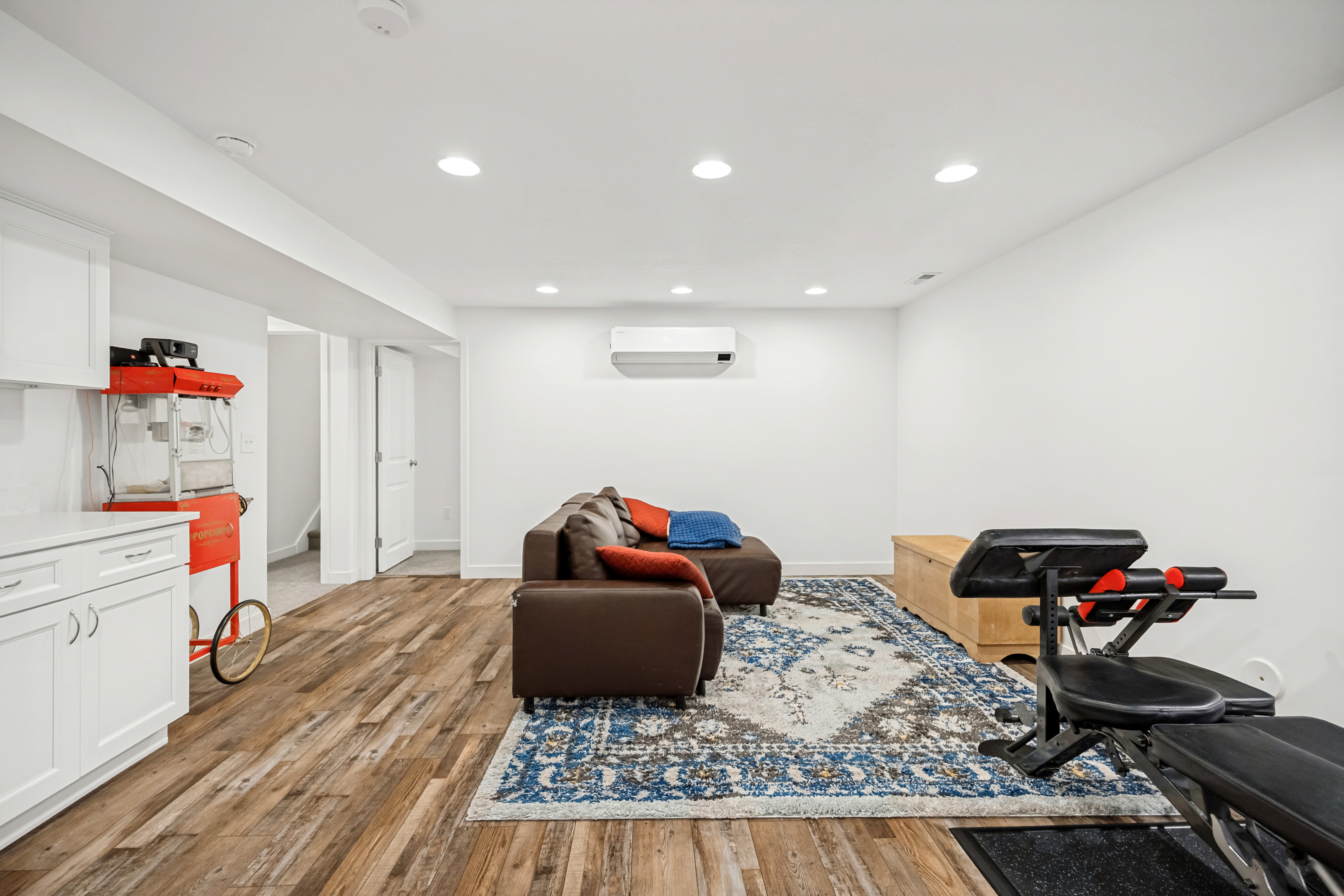 Open-concept recreation area featuring wood-finish flooring, white cabinetry with a light-toned countertop, recessed lighting, and a wall-mounted climate control unit