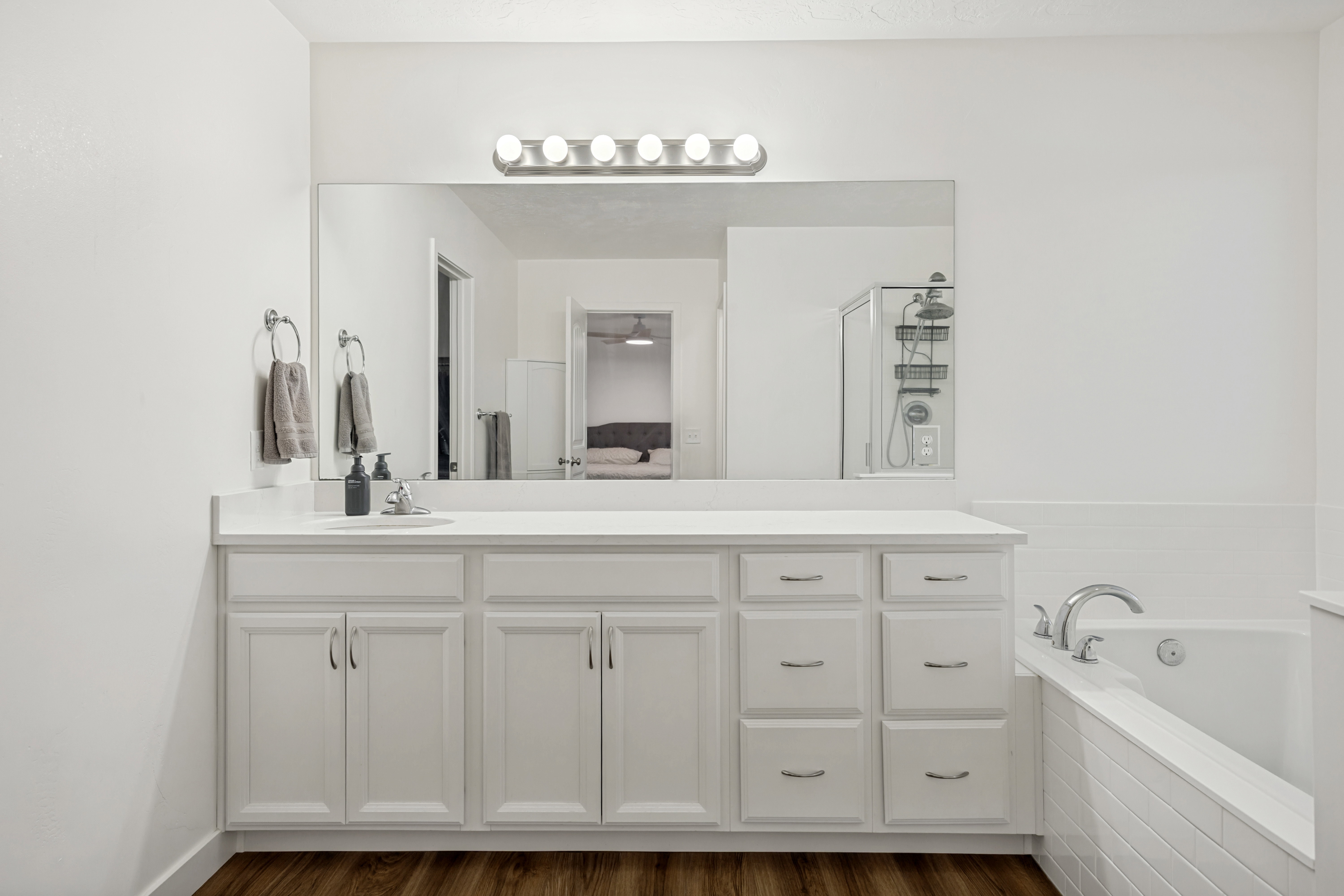 Bathroom featuring a double vanity with white cabinetry and chrome hardware, a large mirror, a chrome light fixture with five bulbs, a built-in bathtub with white tile surround, and wood-finish flooring