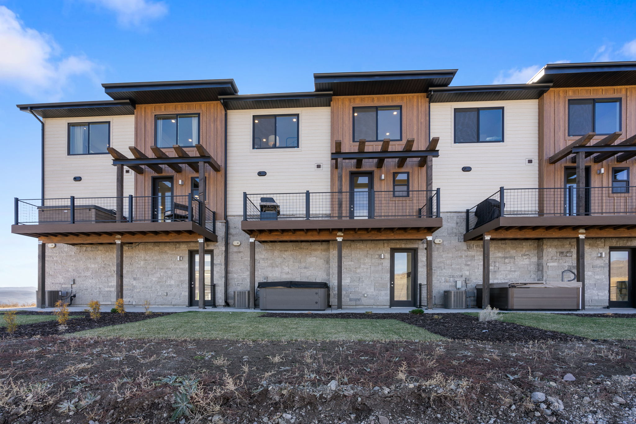 Modern townhome exterior featuring multi-level decks with black railings, wood-look siding accents, and stone veneer foundations