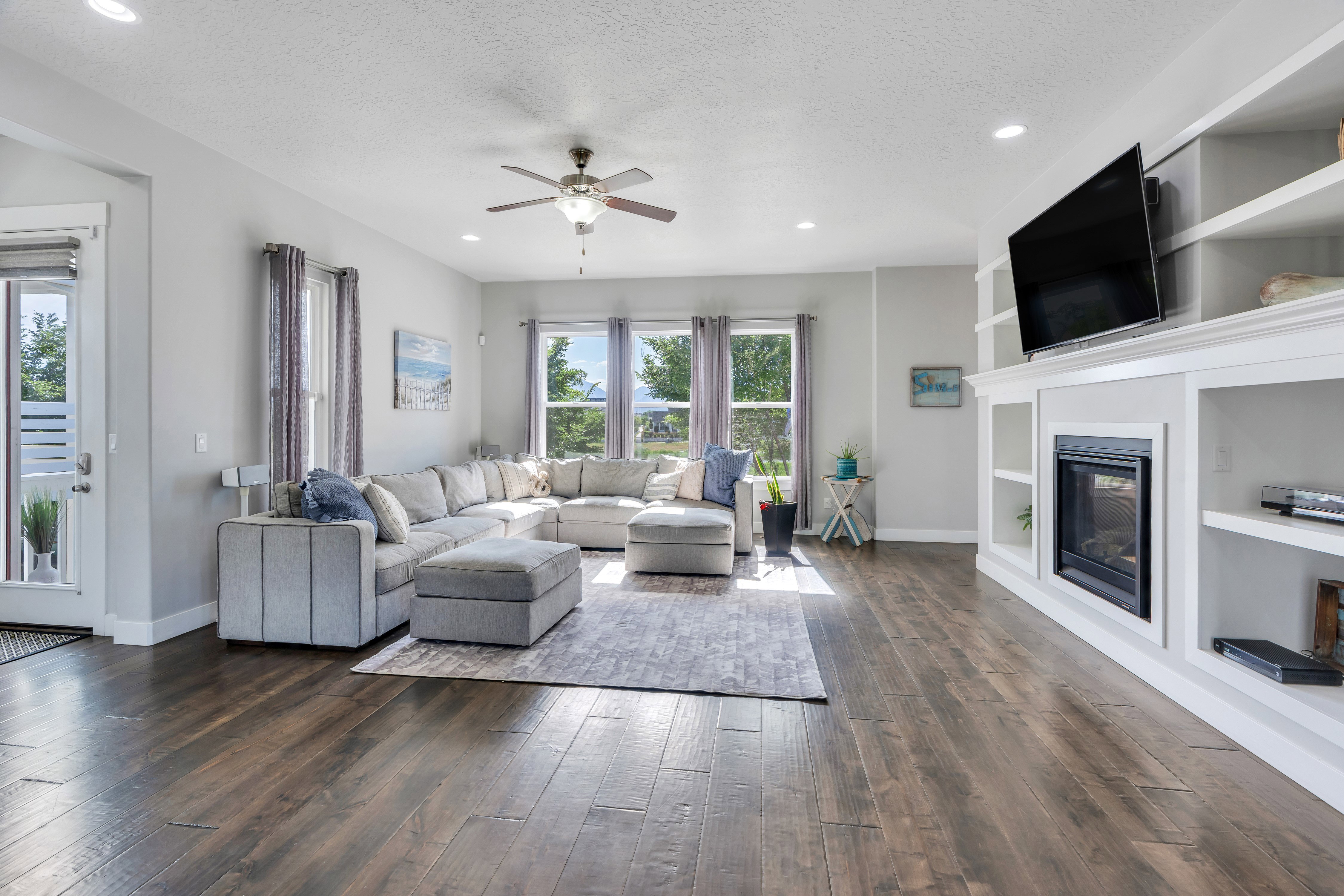 Spacious living area featuring dark wood-finish flooring, a built-in entertainment center with integrated fireplace, recessed lighting, a ceiling fan, and multiple windows providing ample natural light