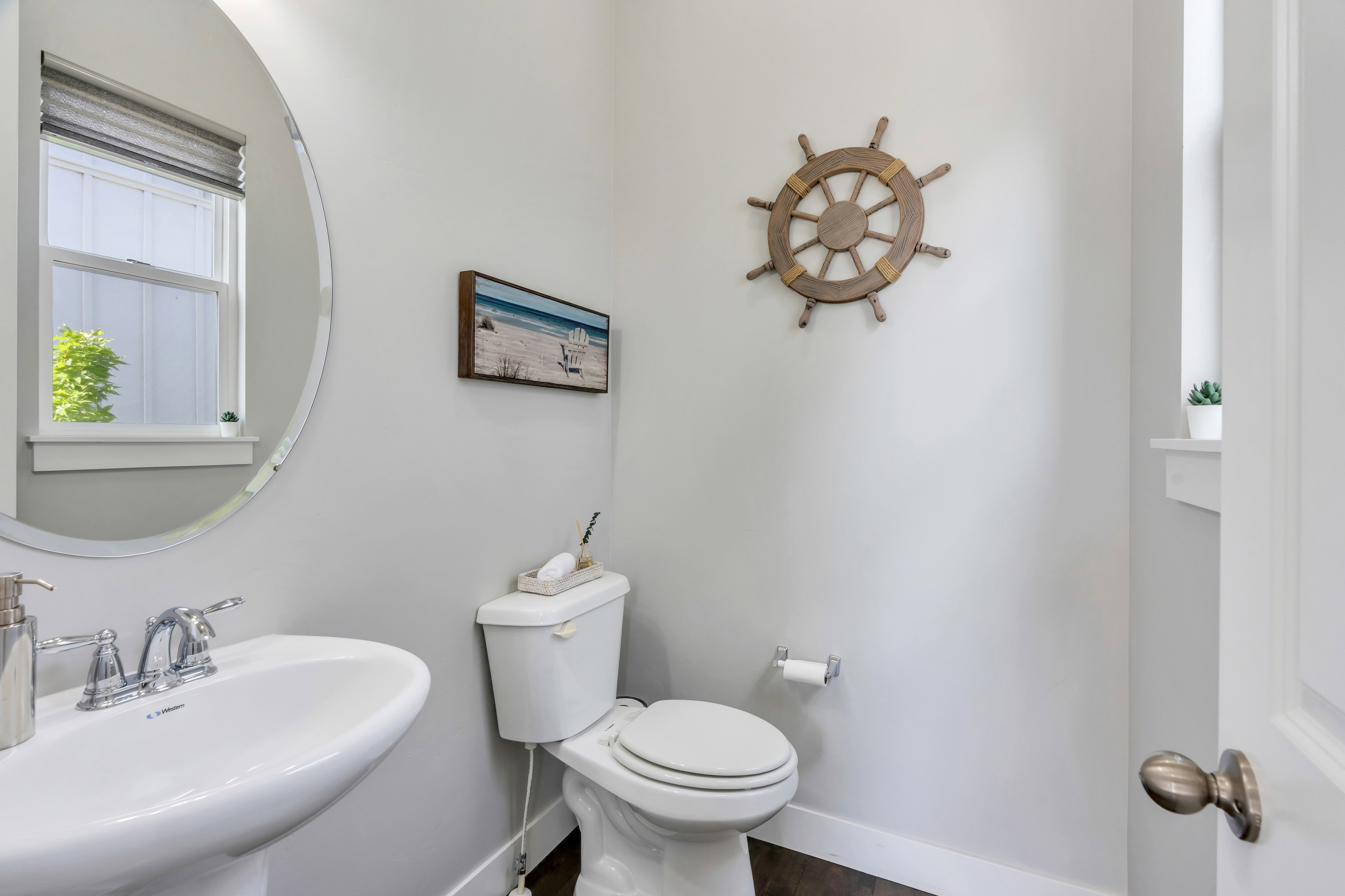 Pedestal sink with chrome faucet, oval mirror, toilet, window with privacy blind, and dark wood-finish flooring